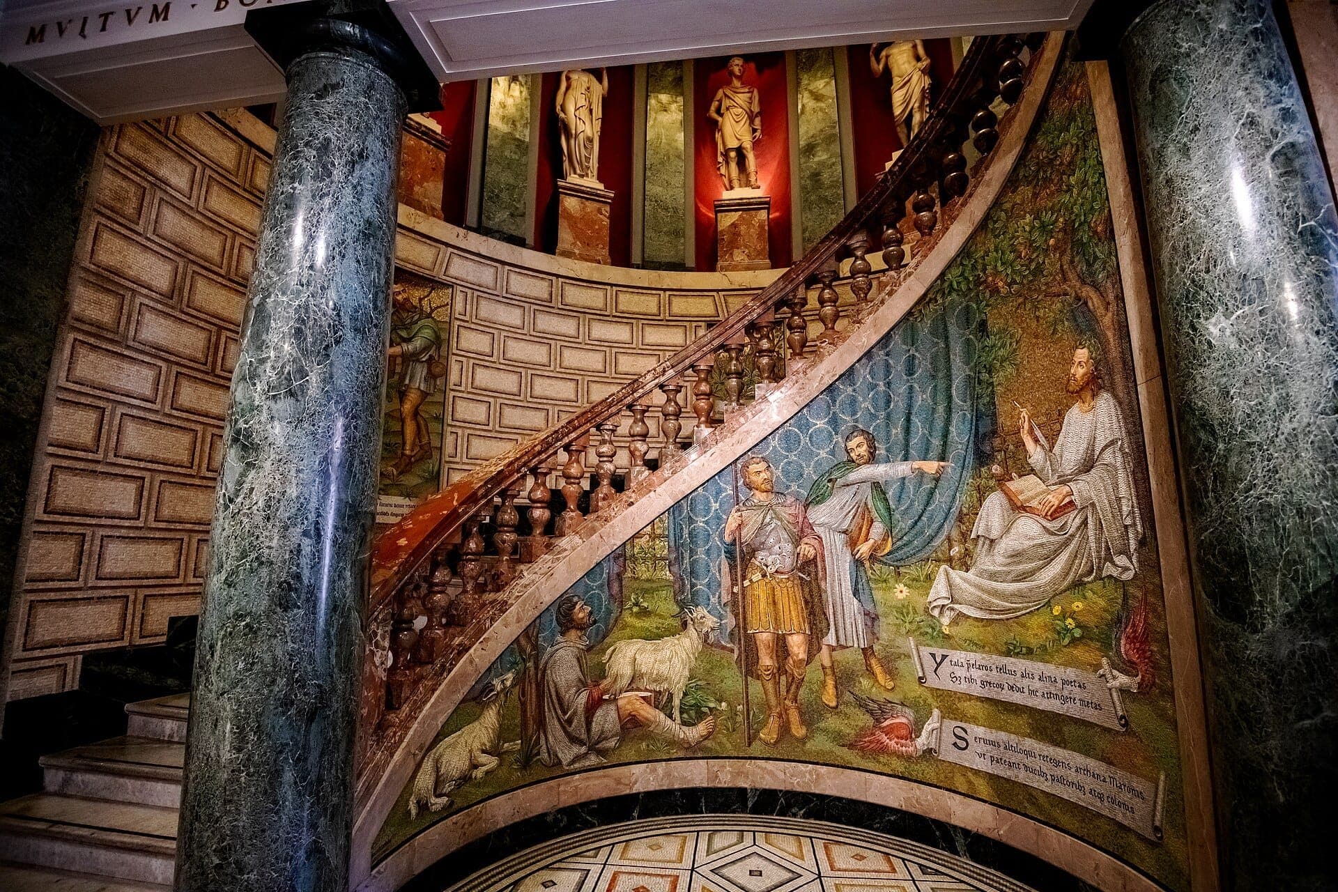 Marble staircase with a vivid mosaic and classical statues at Pinacoteca Ambrosiana in Milan, featuring ornate columns and detailed wall art.