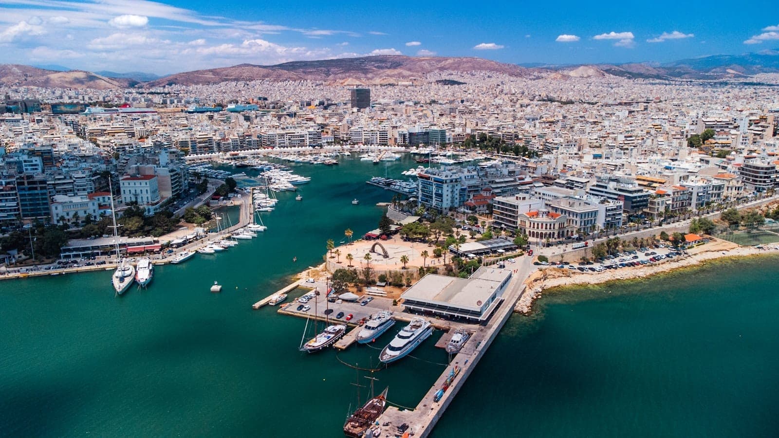 Aerial view of Piraeus port with turquoise water, moored yachts, modern buildings, and hills in the background under a blue sky.