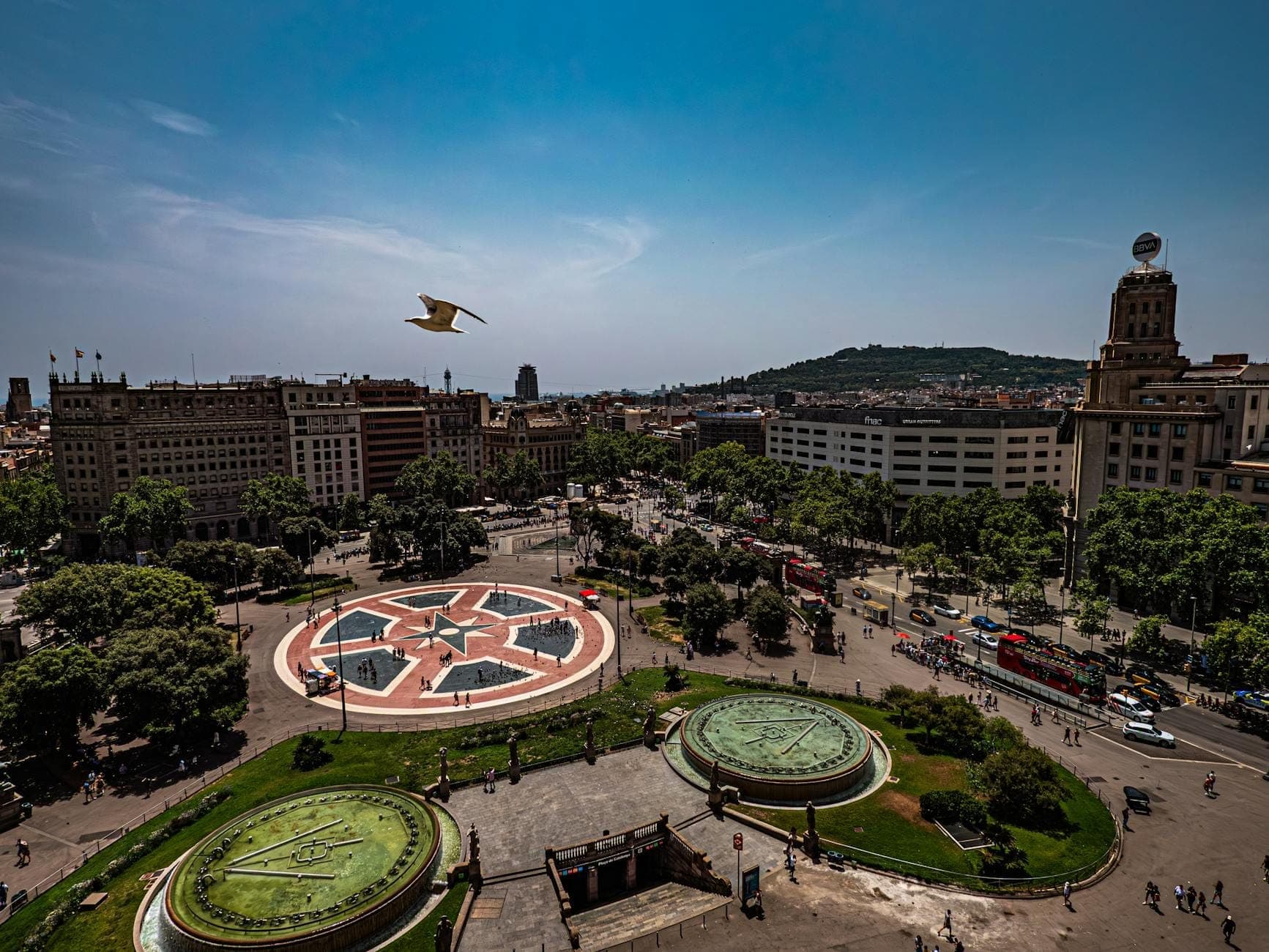 Toàn cảnh Plaça de Catalunya từ trên cao với quảng trường tròn đặc trưng, vườn hoa xung quanh, các tòa nhà lân cận và người dân đang thư giãn tại quảng trường công cộng nhộn nhịp ở Barcelona.