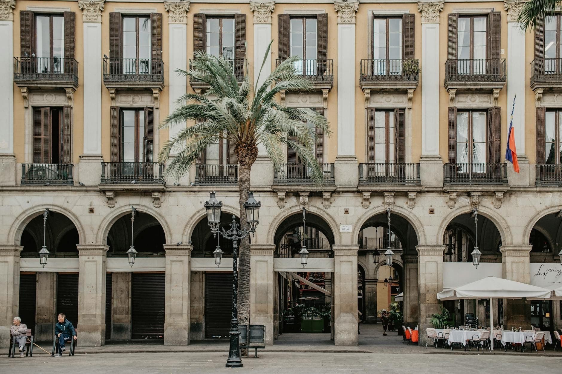 Những tòa nhà tân cổ điển màu vàng với hành lang vòm, ban công và cây cọ tại Plaça Reial, Barcelona — bên dưới là khu vực ăn uống ngoài trời và du khách ngồi thư giãn trong quảng trường.