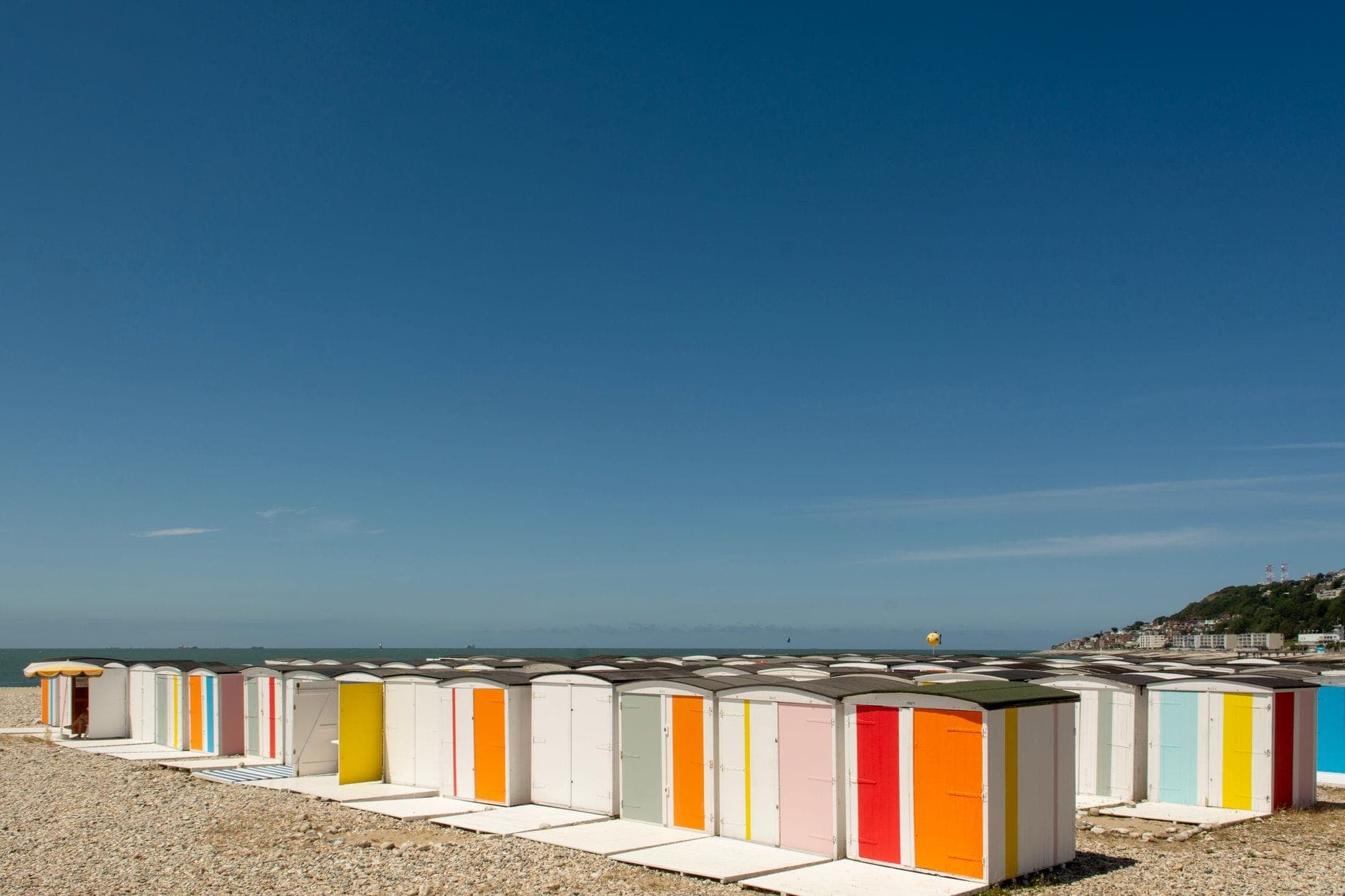 Dãy cabin bãi biển sọc màu nổi bật trên bãi sỏi của Plage du Havre, bầu trời xanh và vách đá xa xa đổ về biển.