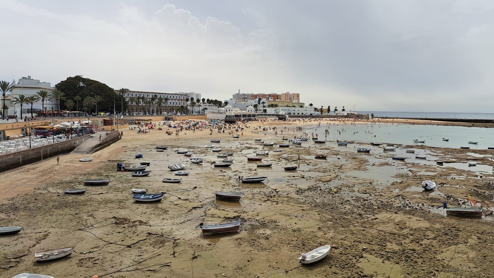 Toàn cảnh Playa de La Caleta lúc thủy triều rút, những chiếc thuyền nhỏ nằm trên cát ướt, phía xa là tường thành cổ, hàng cọ và du khách trên bãi biển.
