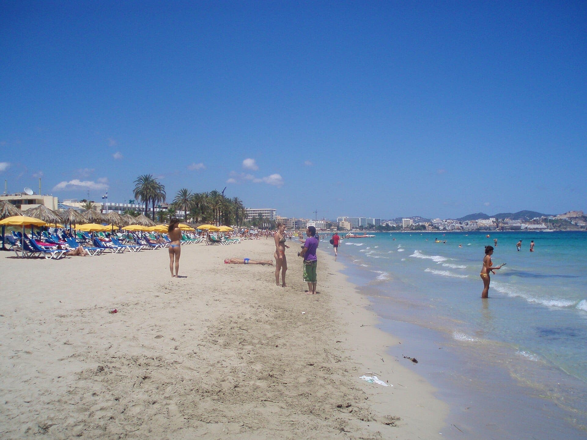 Wide view of Playa d'en Bossa beach with sun loungers, yellow umbrellas, palm trees, and people enjoying the shoreline under clear blue skies.