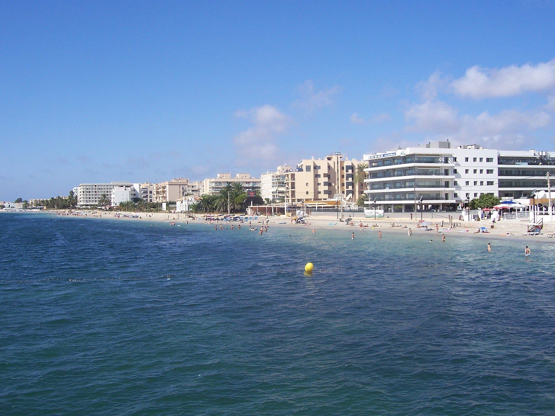 View of Playa d’en Bossa with modern beachfront hotels, sandy shore, sunbathers, and clear blue water under a bright sky.