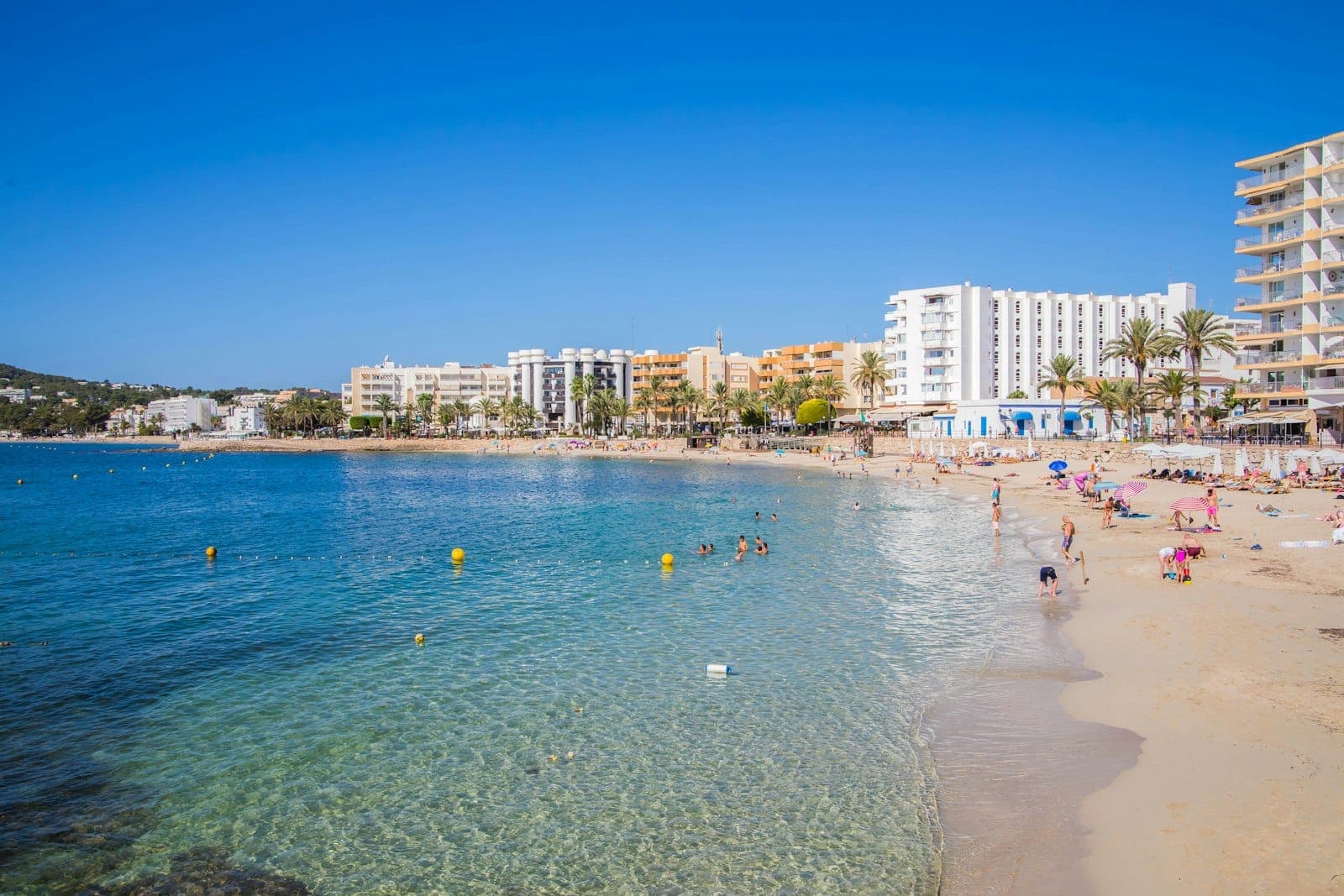 Wide sandy beach lined with resort hotels and palm trees, clear turquoise water with people swimming and relaxing under a bright blue sky.