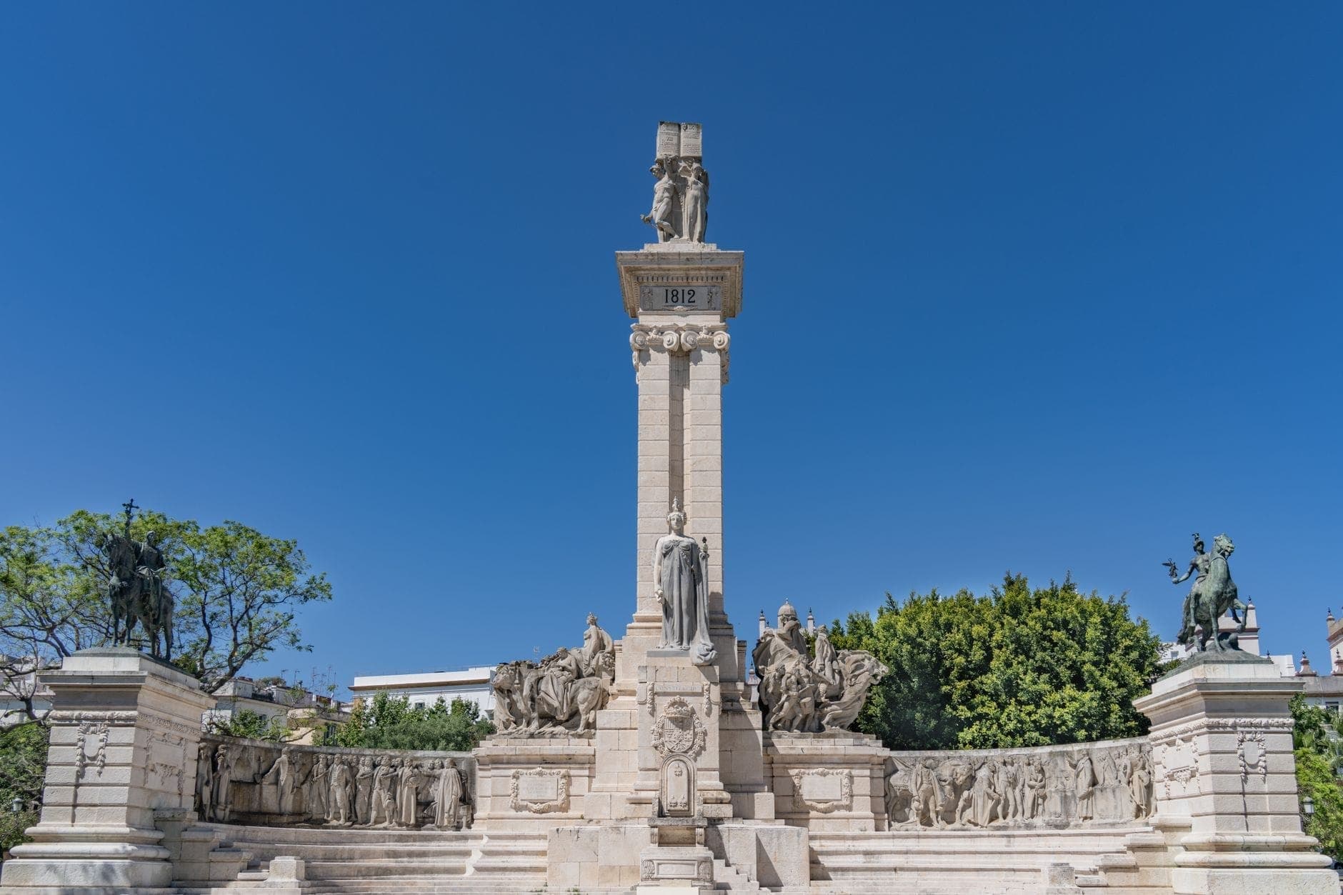 Toàn cảnh Đài tưởng niệm Hiến pháp 1812 tại Plaza de España ở Cádiz, dưới bầu trời xanh và cây xanh bao quanh.