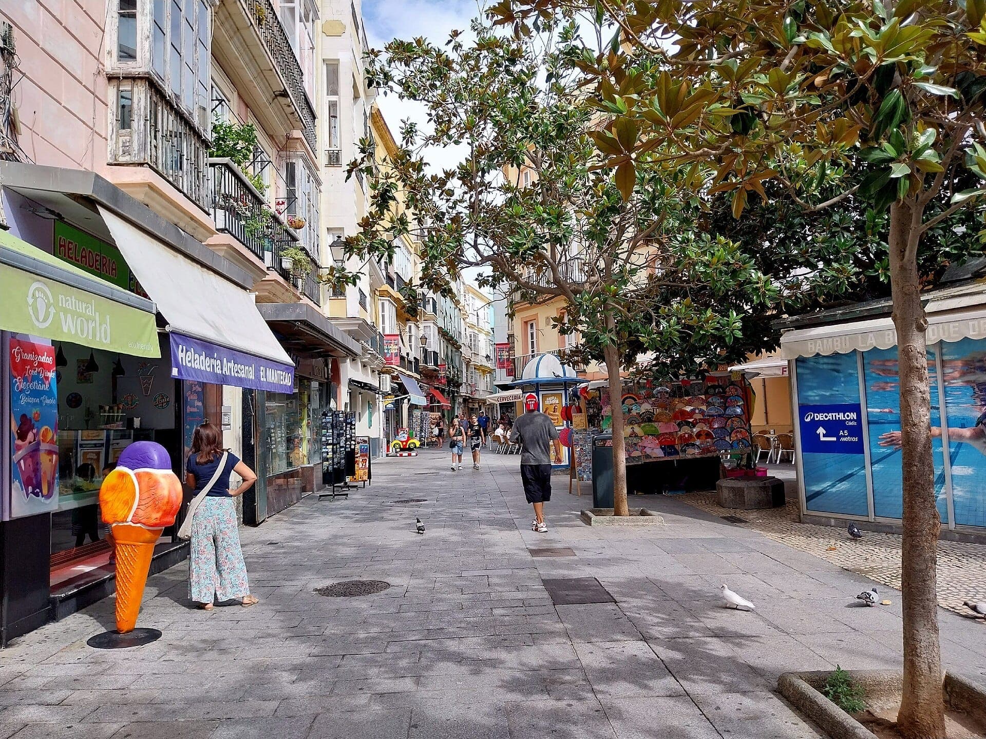 Phố đi bộ tại Plaza de las Flores, Cádiz, với cửa hàng kem đầy màu sắc, quầy bán ô, cây xanh và các tòa nhà lịch sử dưới ánh sáng ban ngày.