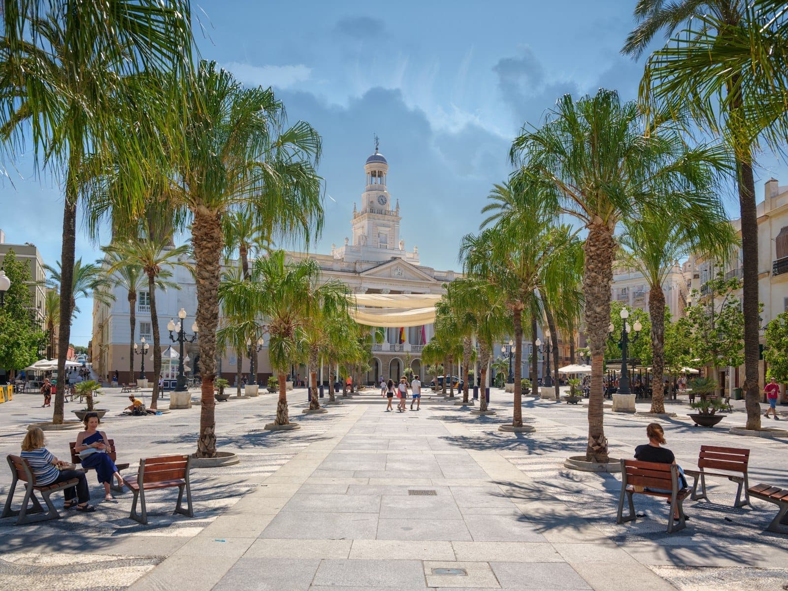 Toàn cảnh Plaza de San Juan de Dios ở Cádiz với hàng cọ, ghế dài, người qua lại và tòa thị chính tân cổ điển phía sau.