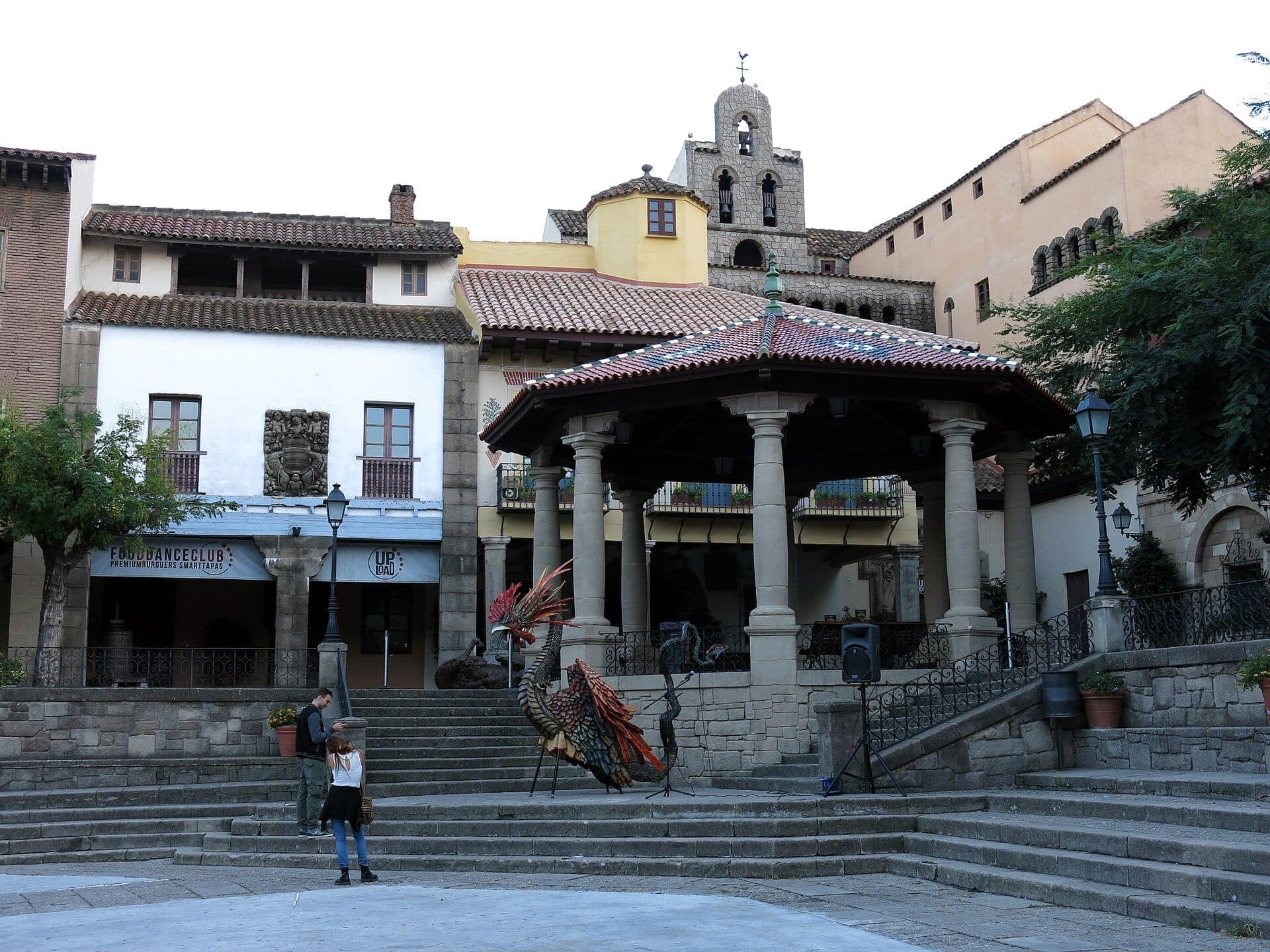 Quảng trường trung tâm tại Poble Espanyol, nổi bật với kiến trúc Tây Ban Nha lịch sử, gian hàng có cột trụ, tượng gà trống ghép gạch mosaic và du khách tham quan vào ban ngày.