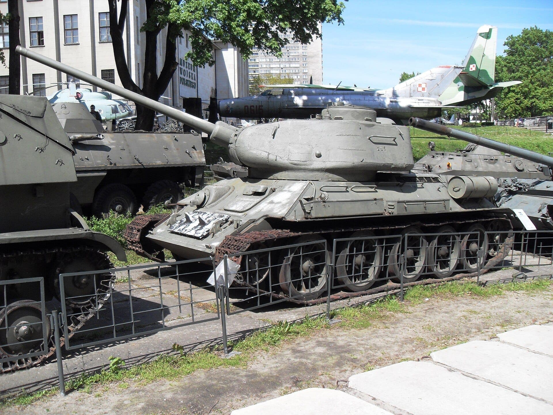 Freiluftausstellung historischer Militärpanzer und Flugzeuge im Polnischen Armeemuseum in Warschau, mit grünen Bäumen und Museumsgebäuden im Hintergrund.