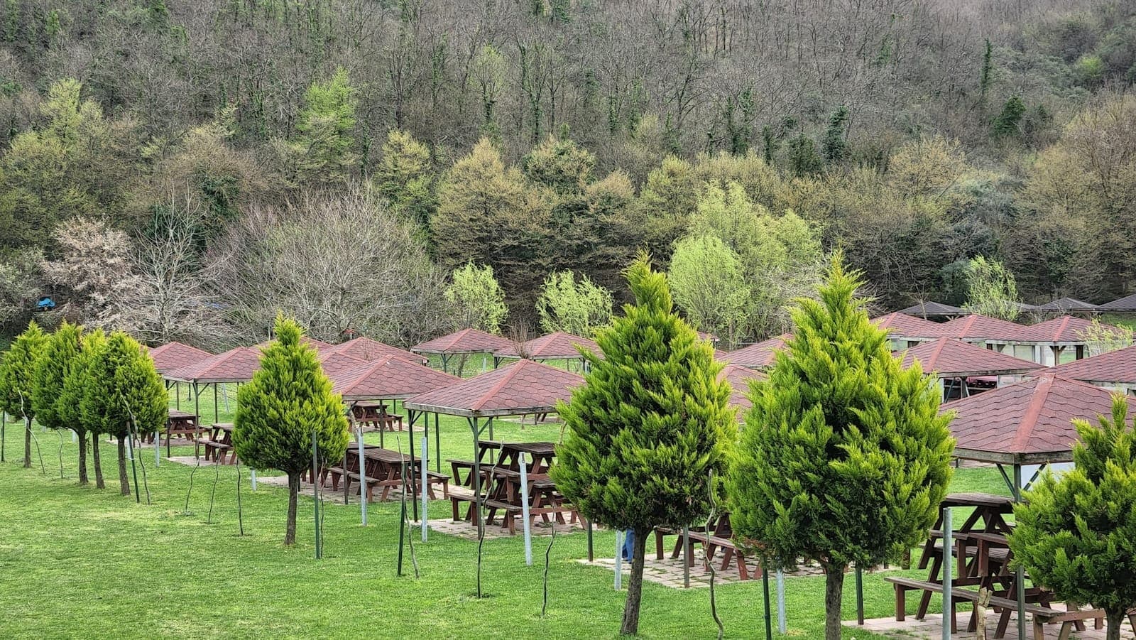 Reihen von überdachten Picknicktischen und Holzpavillons im Naturpark Polonezköy, umgeben von grünen Bäumen und einem bewaldeten Hang.