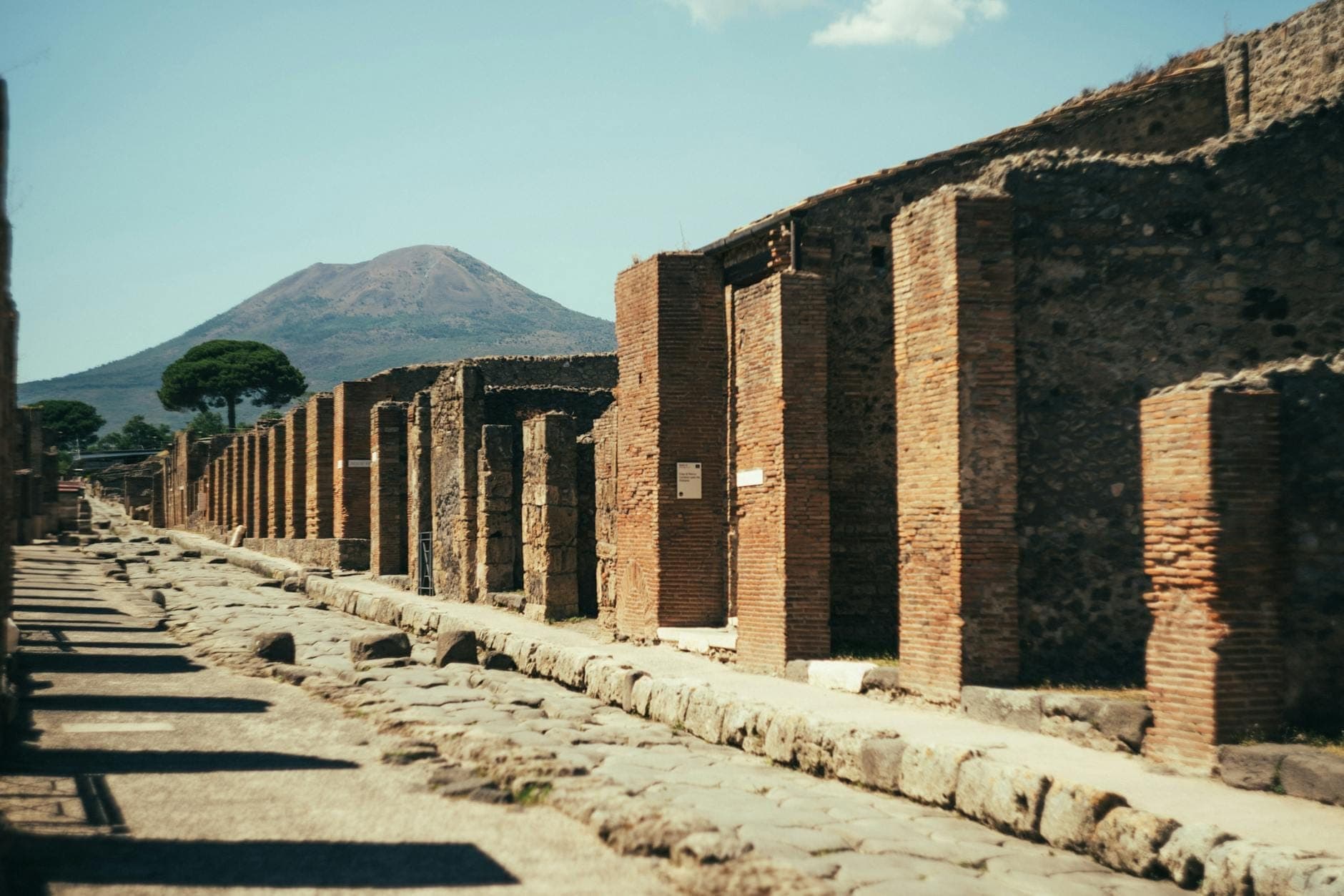 Cảnh phế tích Pompeii với những con đường lát đá, tường gạch cổ và núi Vesuvius in bóng phía sau dưới bầu trời xanh trong.