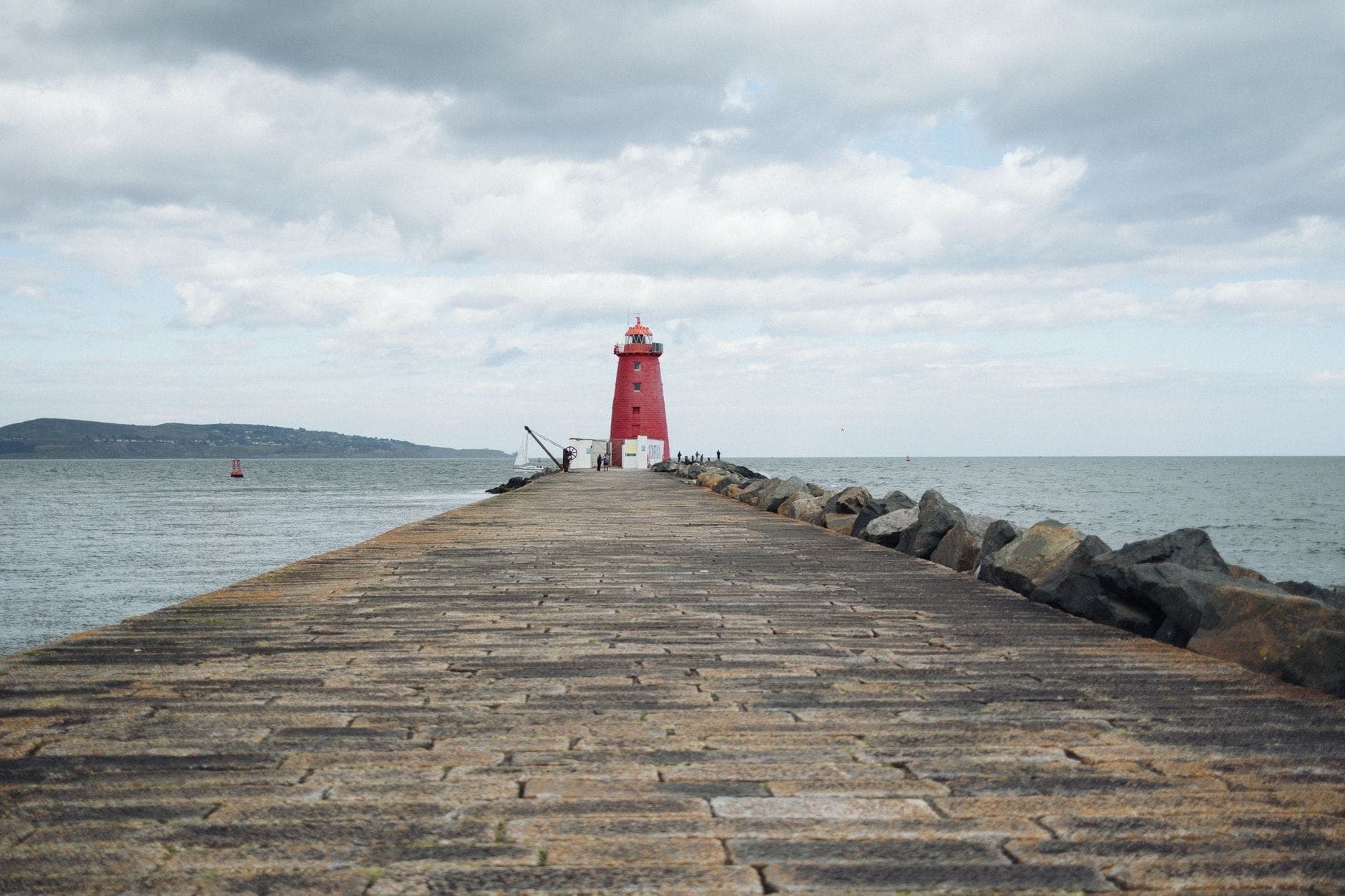 Blick entlang der steinernen Great South Wall, die direkt auf den leuchtend roten Poolbeg Lighthouse zuführt — auf beiden Seiten das Meer, darüber ein bewölkter Himmel.