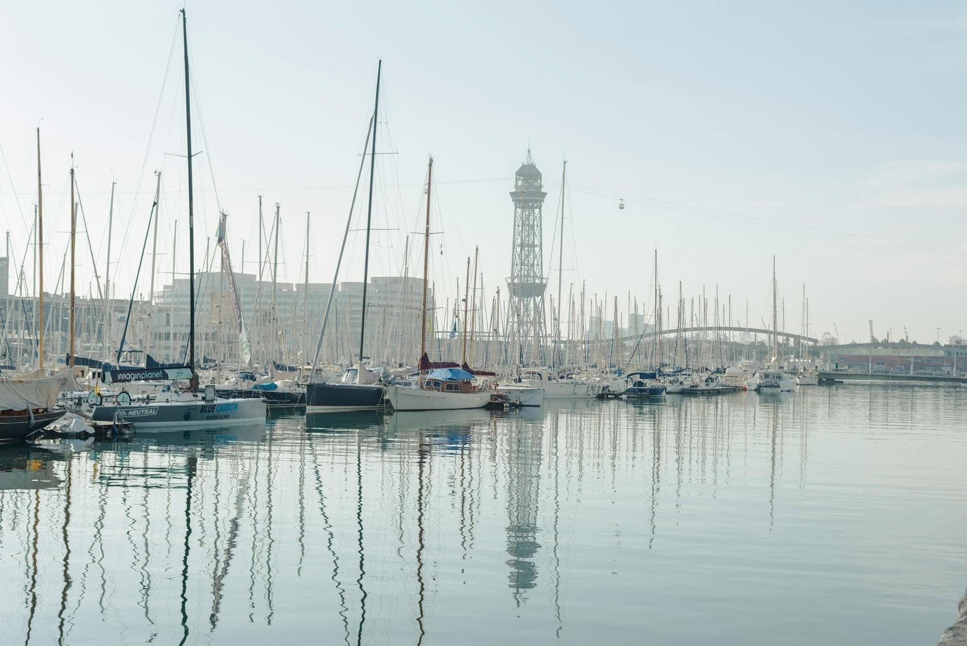 Toàn cảnh bến cảng Port Vell ở Barcelona với những chiếc thuyền buồm neo đậu, mặt nước phẳng lặng và tháp cáp treo biểu tượng hiện ra phía xa dưới bầu trời trong xanh.