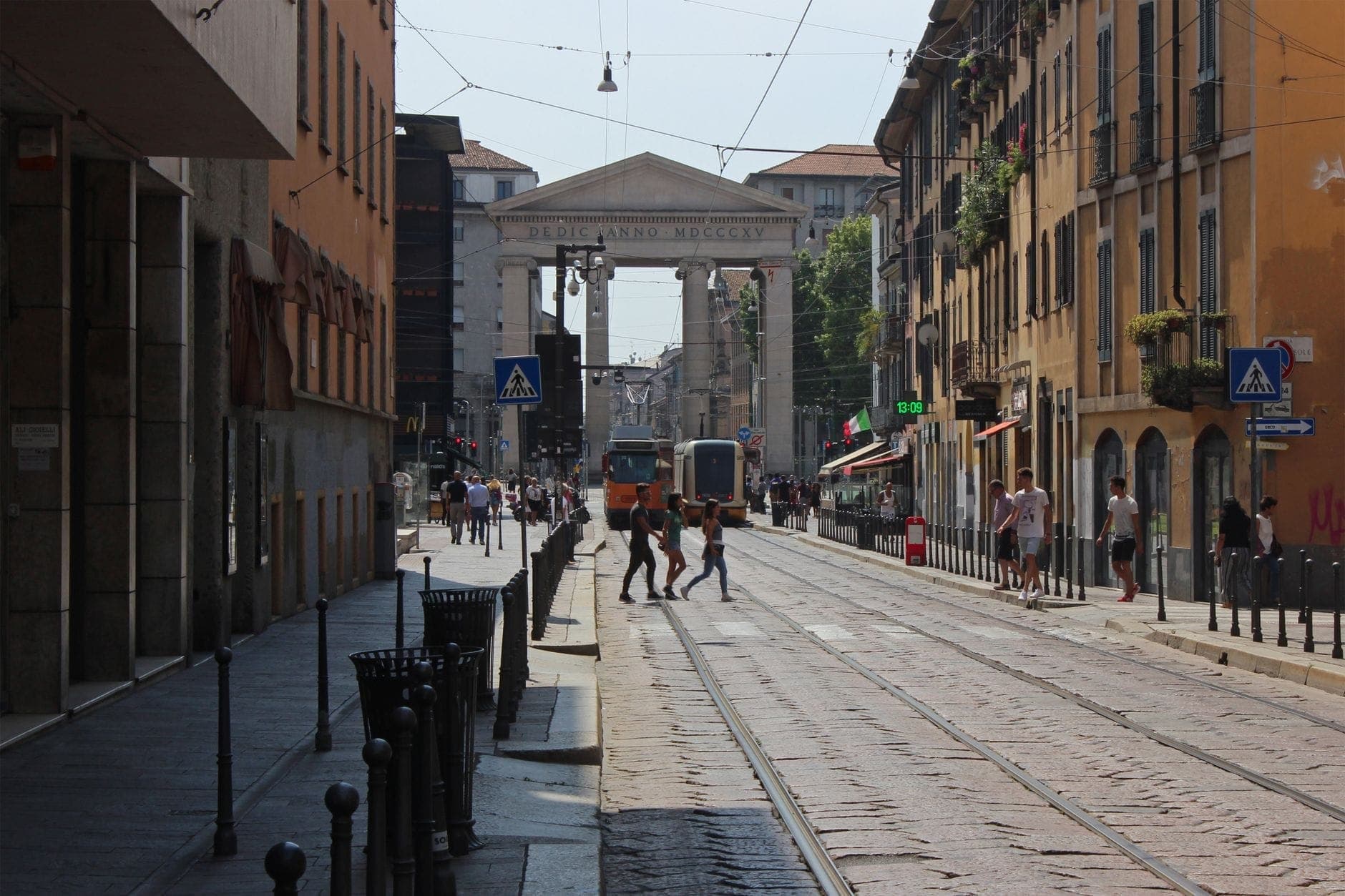 Street view of Porta Ticinese in Milan with people crossing tram tracks, neoclassical gate visible at the end, framed by historic buildings.