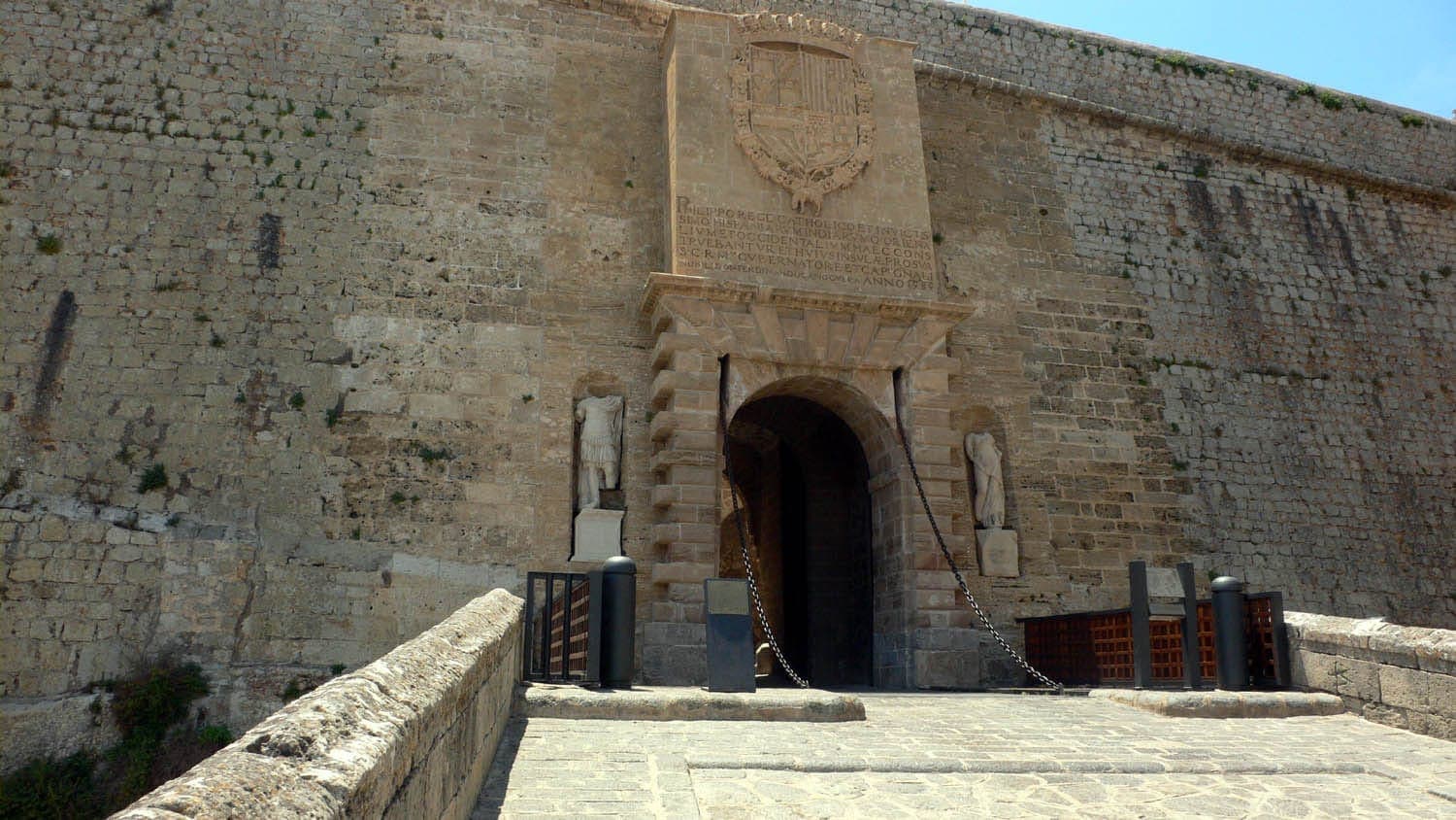 Stone entrance archway of Portal de ses Taules in Ibiza, flanked by statues and ancient walls under sunny blue sky.