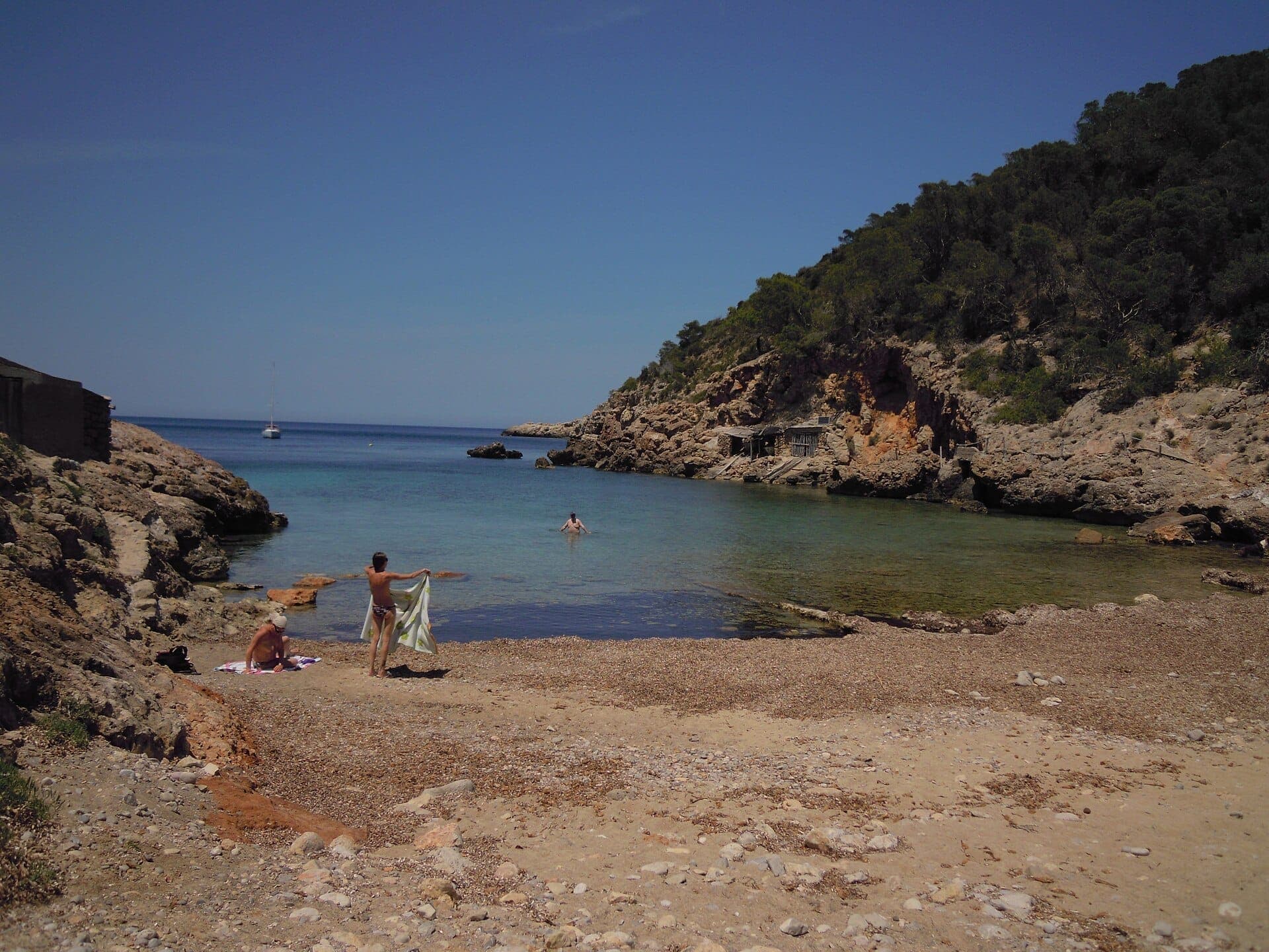 Secluded cove at Portinatx with calm clear water, rocky cliffs, rustic boat sheds, and a few people enjoying the quiet beach.