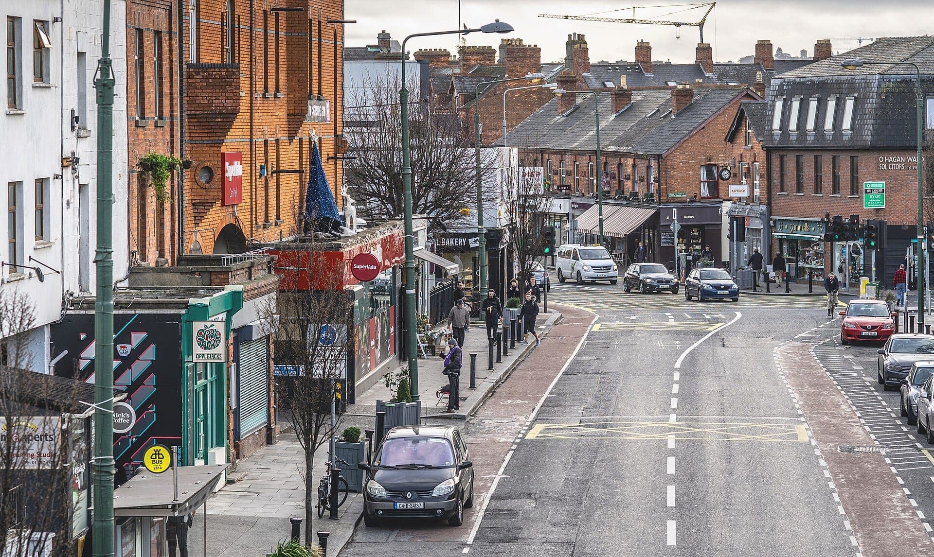 Straßenansicht von Ranelagh in Dublin mit Backsteingebäuden, lokalen Geschäften, geparkten Autos und Menschen, die an einem bewölkten Tag spazieren gehen.