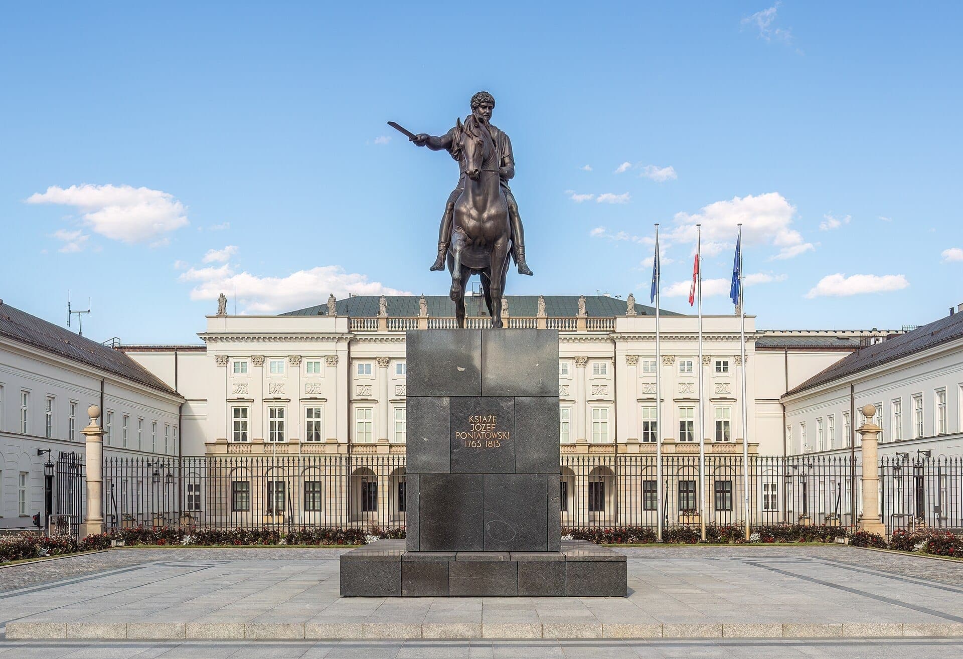 Vorderansicht des Präsidentenpalastes Warschau mit dem Reiterstandbild von Fürst Józef Poniatowski und drei Fahnenmasten an einem klaren Tag.