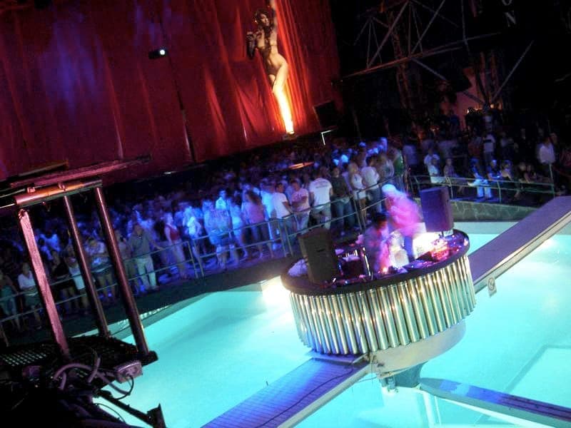 Large crowd inside Privilege Ibiza nightclub, illuminated dance floor and DJ booth over a pool, with dramatic red drapery in the background.