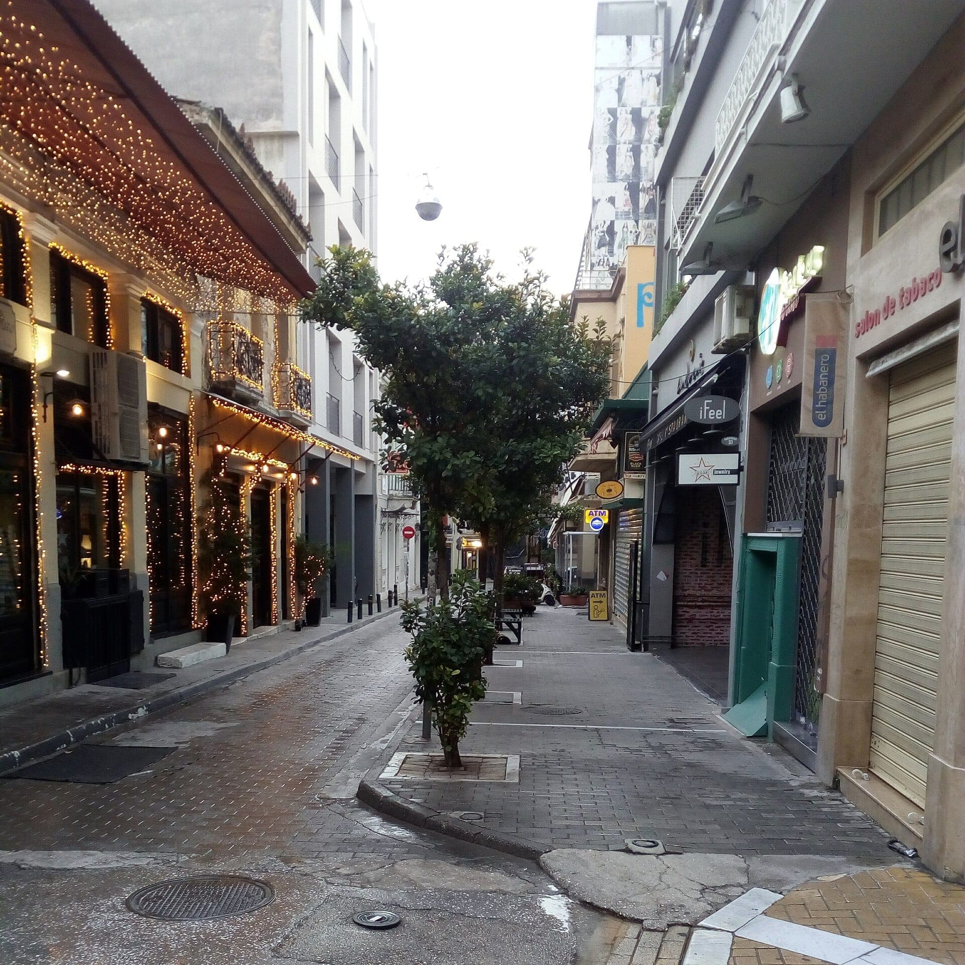 Enge Straße in Psyrri, Athen, mit dekorierten neoklassizistischen Gebäuden, schimmernden Lichterketten und einem baumgesäumten Gehweg an einem ruhigen Tag.