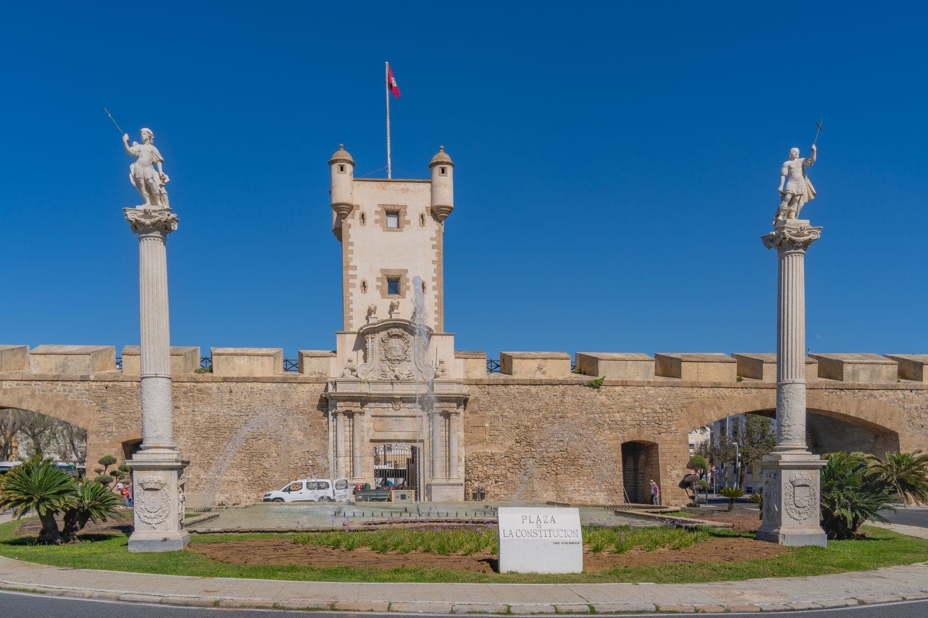 Nhìn thẳng mặt trước Puertas de Tierra tại Cádiz, nổi bật với tượng trên cột, tháp trung tâm và bầu trời xanh trong.