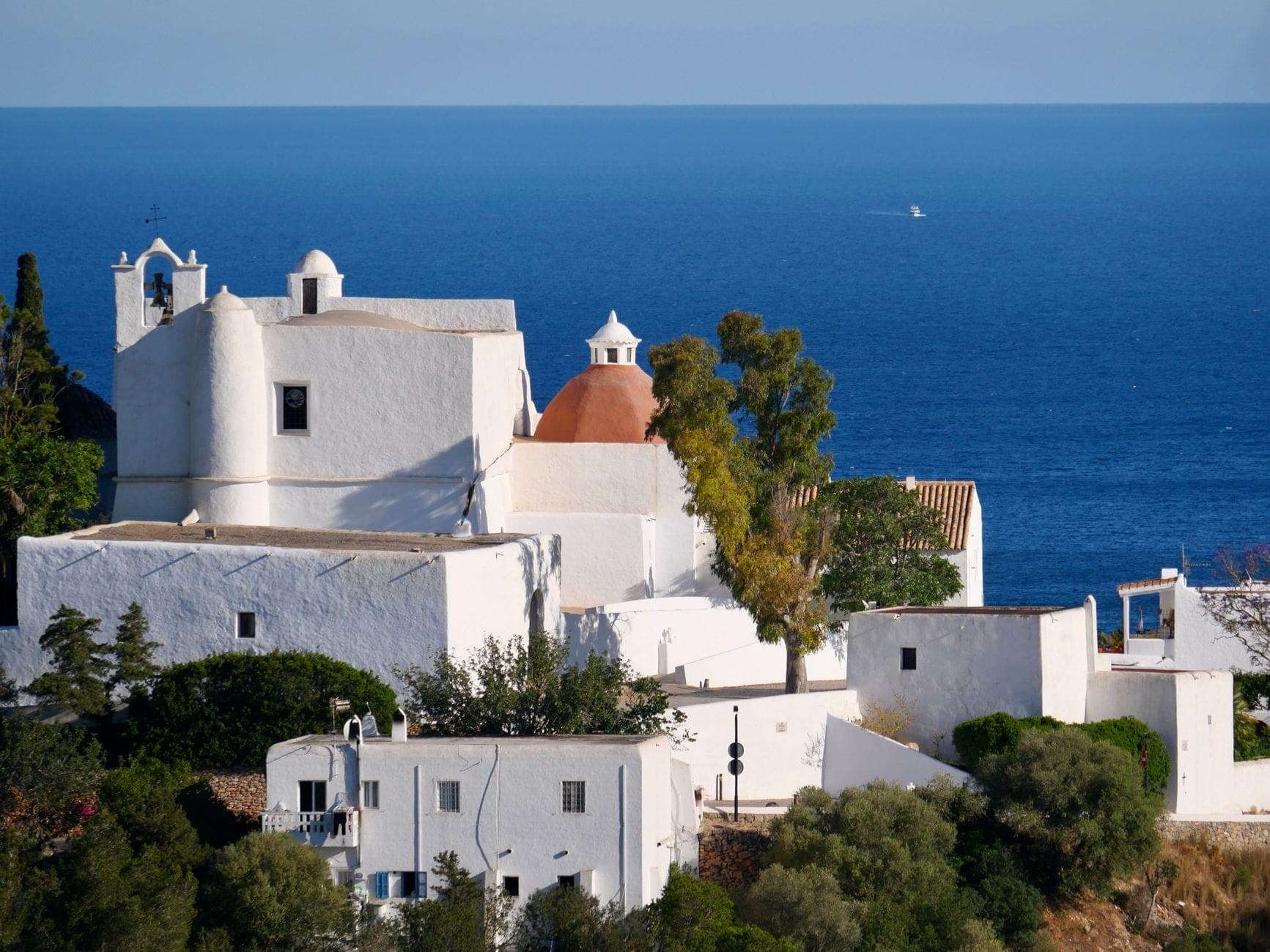 Whitewashed church of Puig de Missa perched on a hilltop overlooking the sea, surrounded by trees and historic buildings in Santa Eulalia, Ibiza.