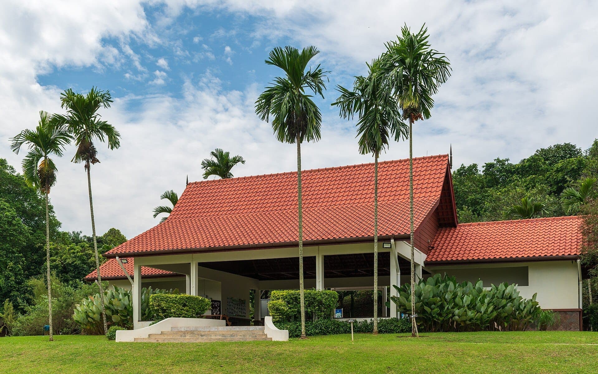 Nhà triển lãm Dân tộc Thực vật học mái đỏ được bao quanh bởi cây nhiệt đới xanh tươi và cây cọ tại Vườn Thực vật Putrajaya dưới bầu trời có mây một phần.