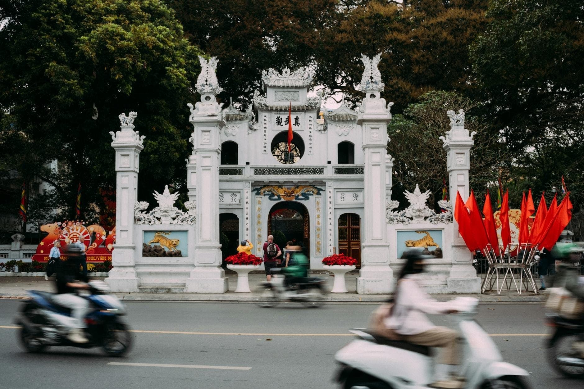 Cổng tam quan trắng tinh xảo của Đền Quán Thánh nhìn từ phía trước, với du khách và xe máy qua lại, nổi bật trên nền cây xanh và cờ đỏ rực rỡ giữa lòng Hà Nội.