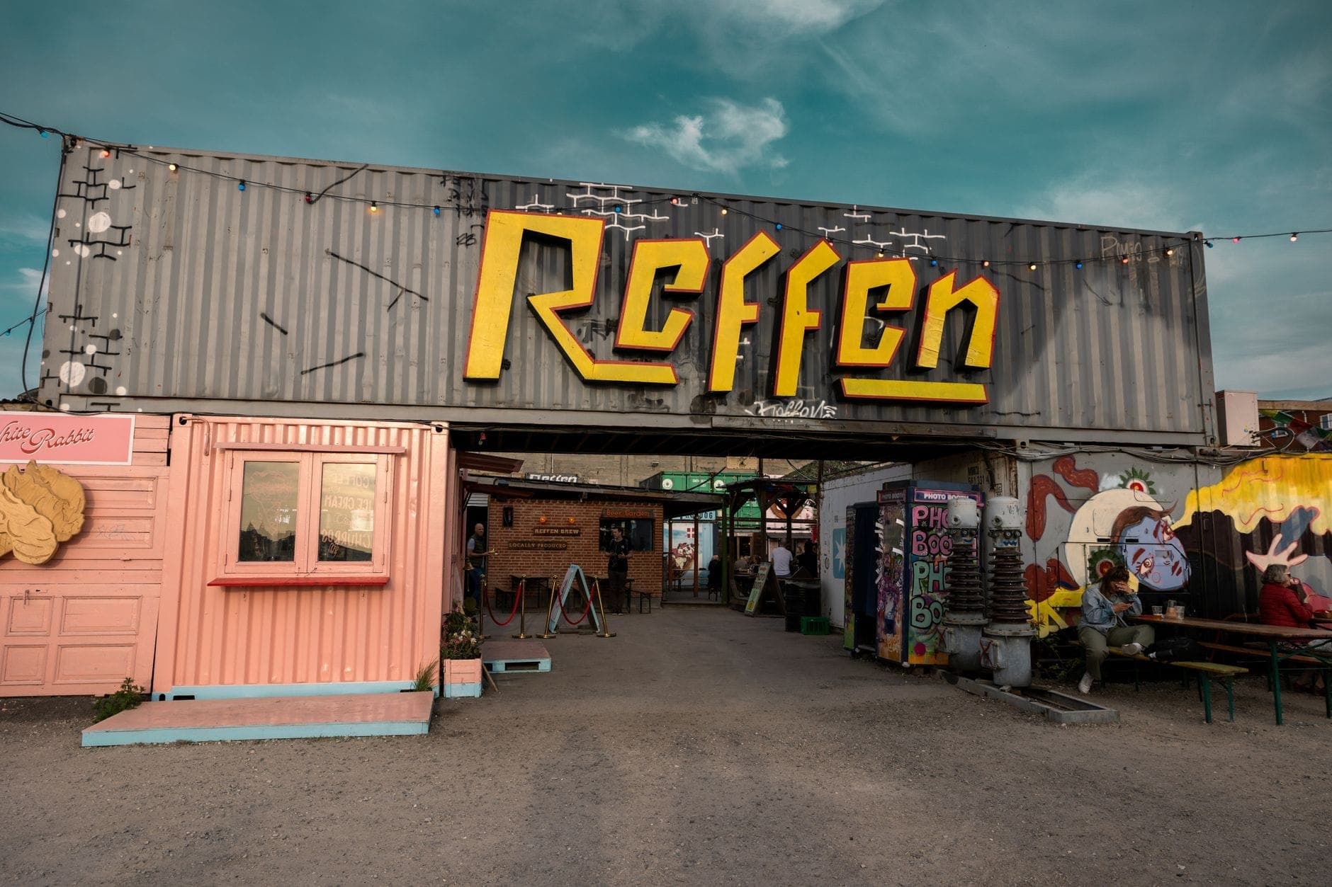 Blick auf den Haupteingang des Streetfood-Markts Reffen mit leuchtendem gelbem Schild, bunten Schiffscontainern und Außenbestuhlung an einem sonnigen Tag.