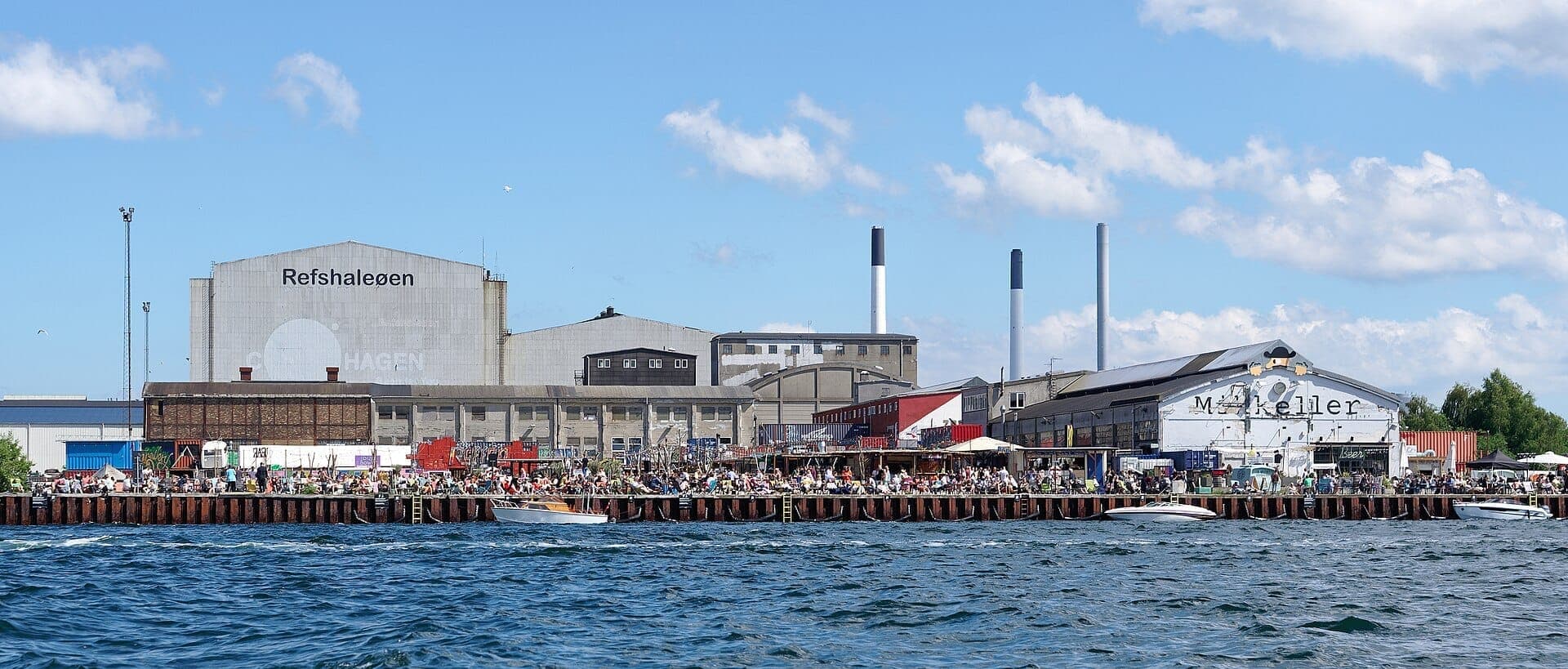 Industriegebäude und Uferpromenade auf Refshaleøen in Kopenhagen, mit vielen Menschen, die einen sonnigen Tag am Hafen genießen.
