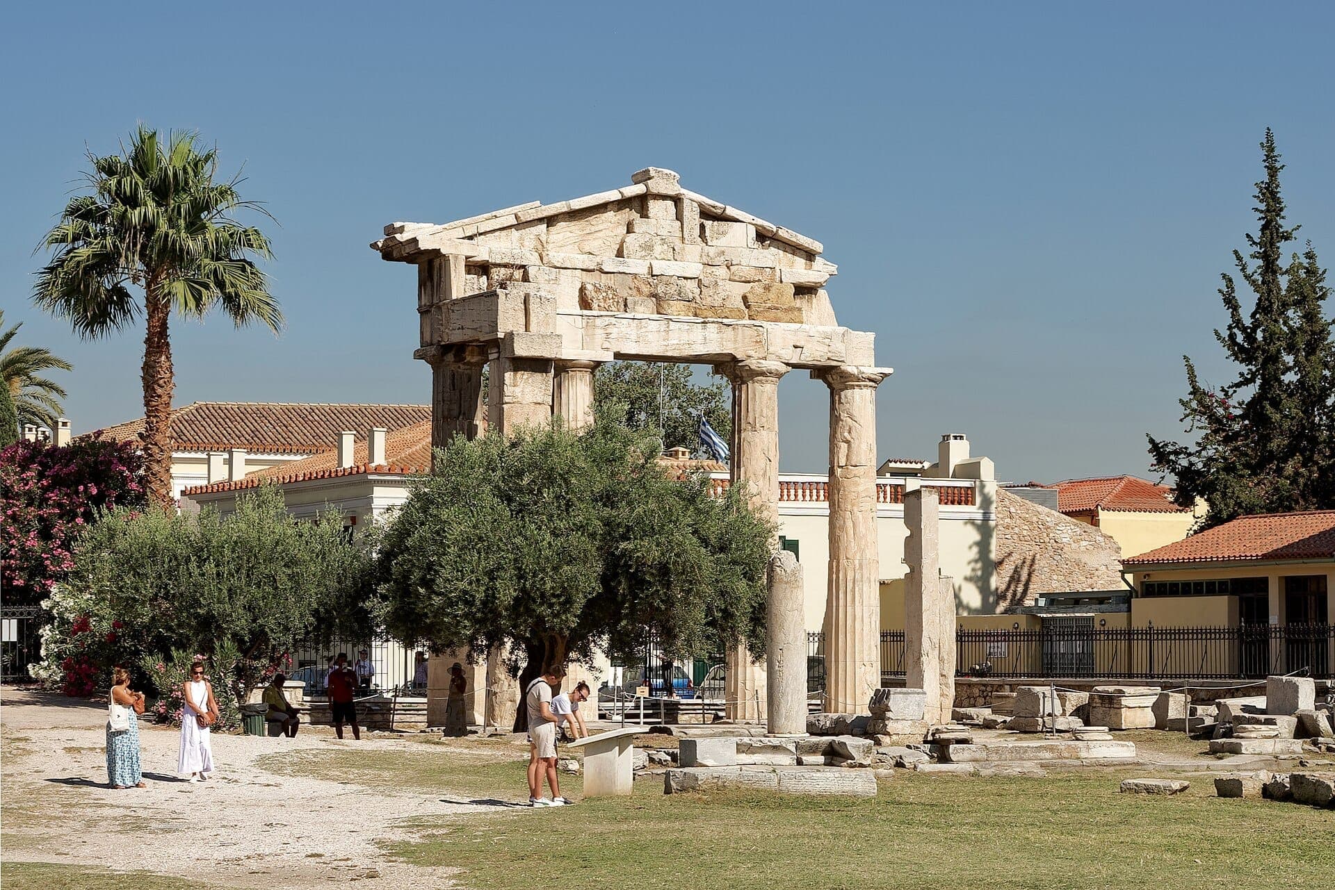 The Gate of Athena Archegetis at the Roman Agora of Athens, surrounded by olive trees, palm trees, and visitors under clear blue skies.