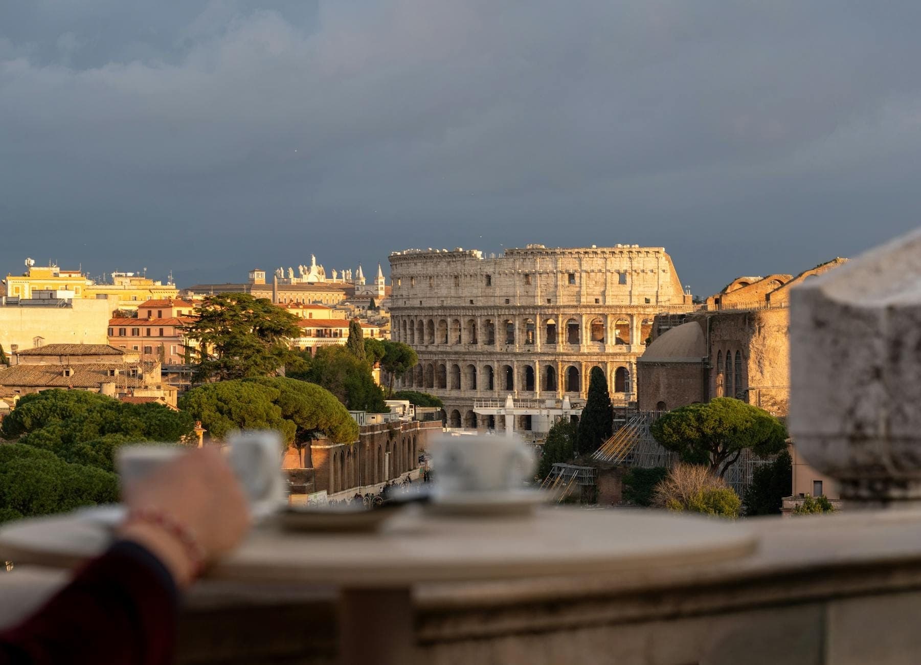 Toàn cảnh Rome với đấu trường Colosseum phía xa và một bàn tay đang cầm tách cà phê ở tiền cảnh dưới nắng vàng.
