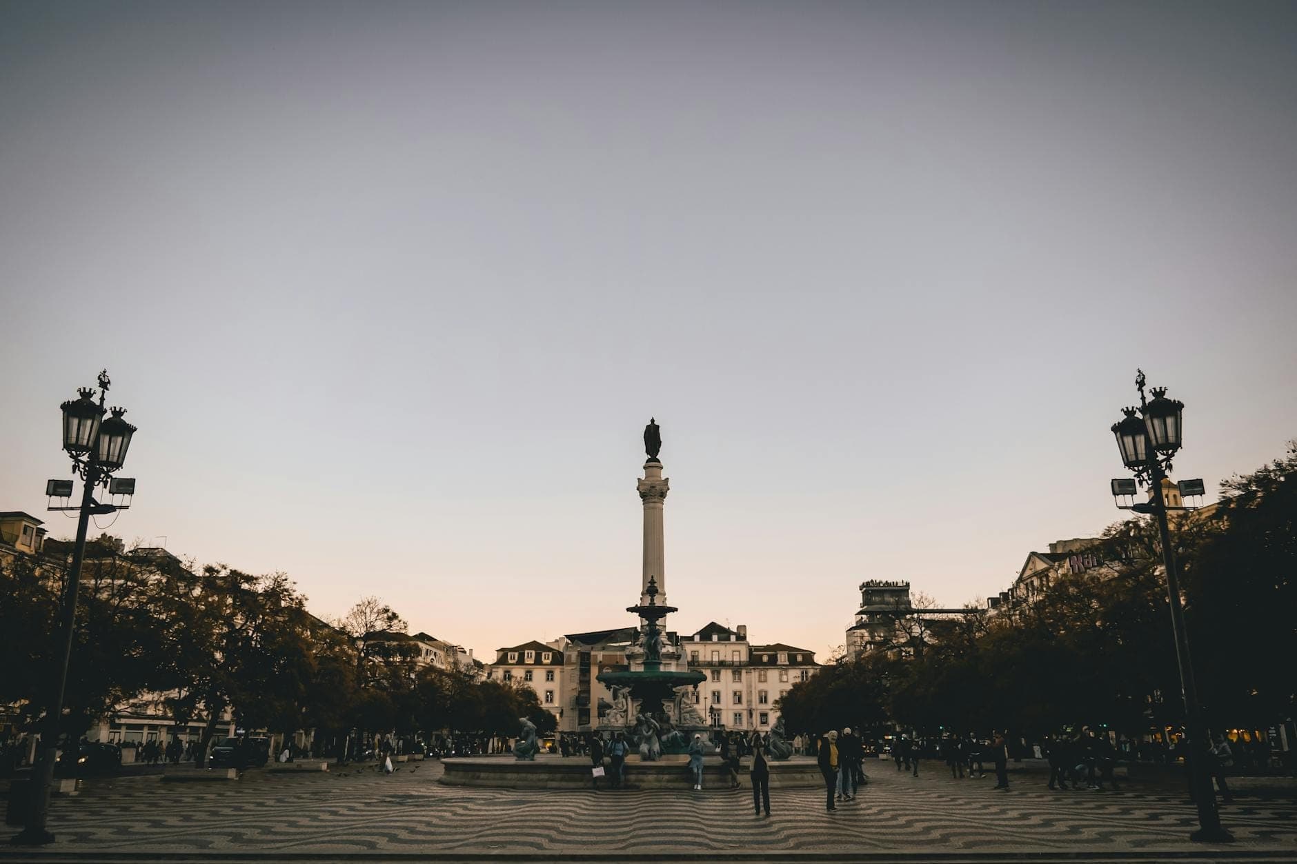 Toàn cảnh Quảng trường Rossio lúc hoàng hôn với cột trung tâm, nền đá hoa văn sóng nước, những tòa nhà cổ kính, đèn đường và người dân tụ họp tại Lisbon, Bồ Đào Nha.