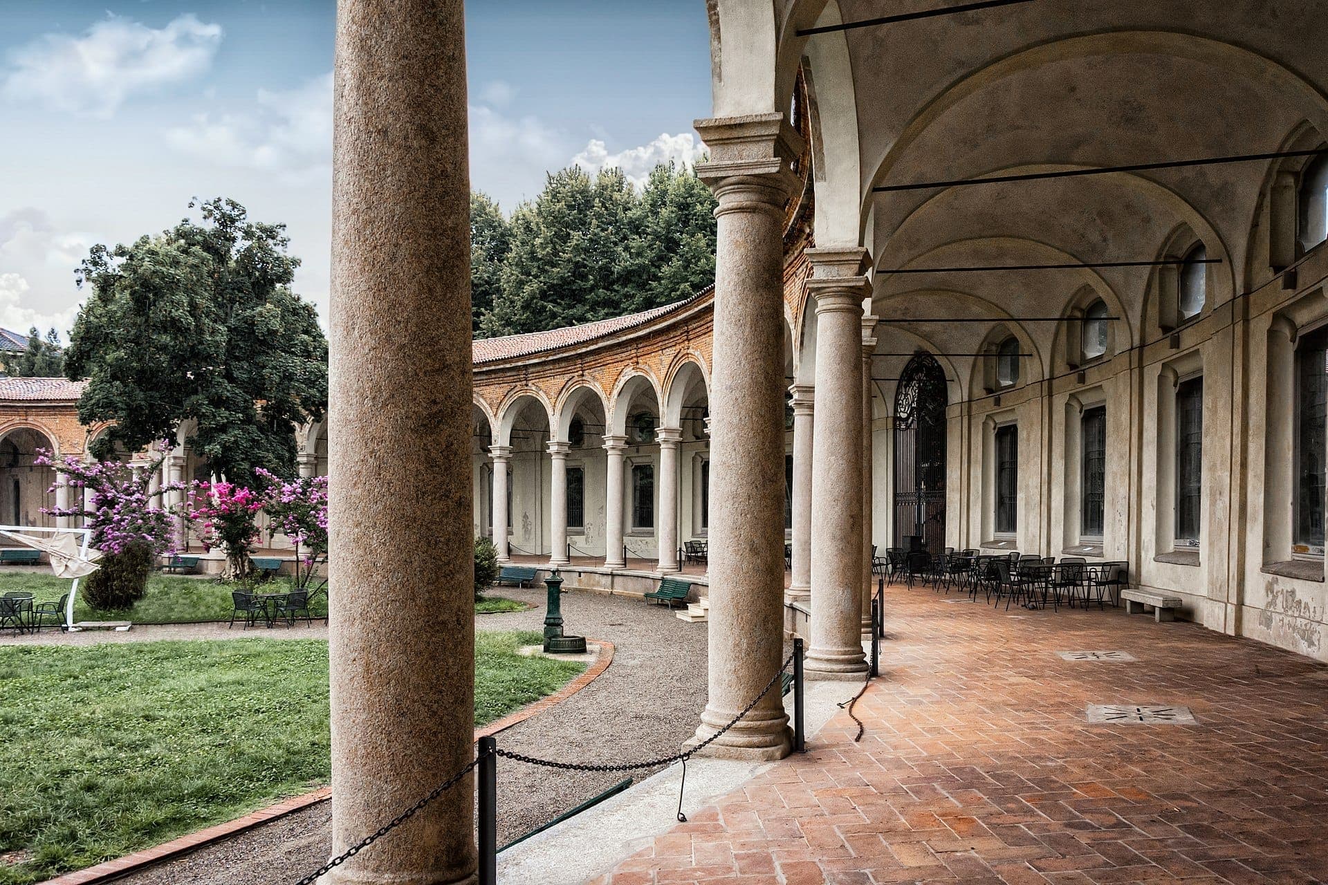 View of Rotonda della Besana’s arched colonnade and tranquil central garden with vibrant flowers and empty outdoor chairs on a cloudy day.