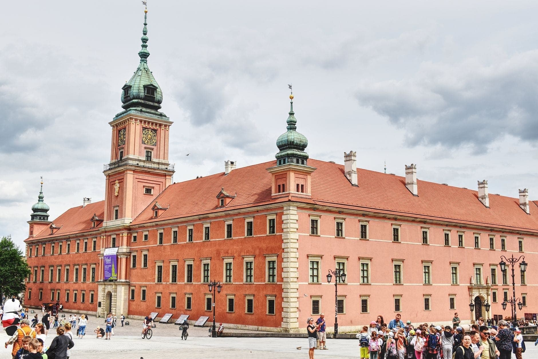 Weitläufige Panoramaansicht des Königsschlosses in Warschau mit seiner imposanten rotbacksteinernen Fassade, den Türmen und dem belebten Schlossplatz voller Besucher unter bewölktem Himmel.