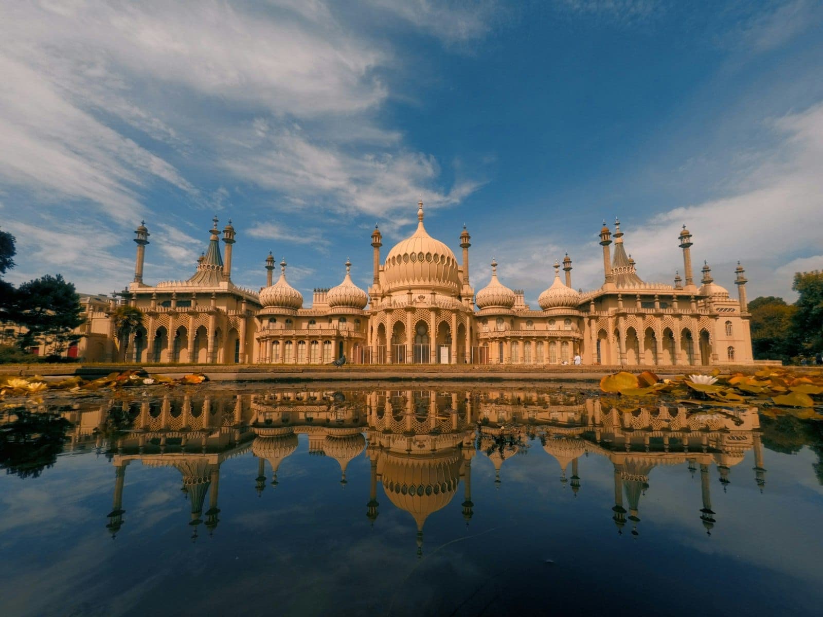 Toàn cảnh Royal Pavilion ở Brighton với mái vòm và tháp nhỏ phản chiếu trên mặt hồ, dưới bầu trời xanh rực rỡ.