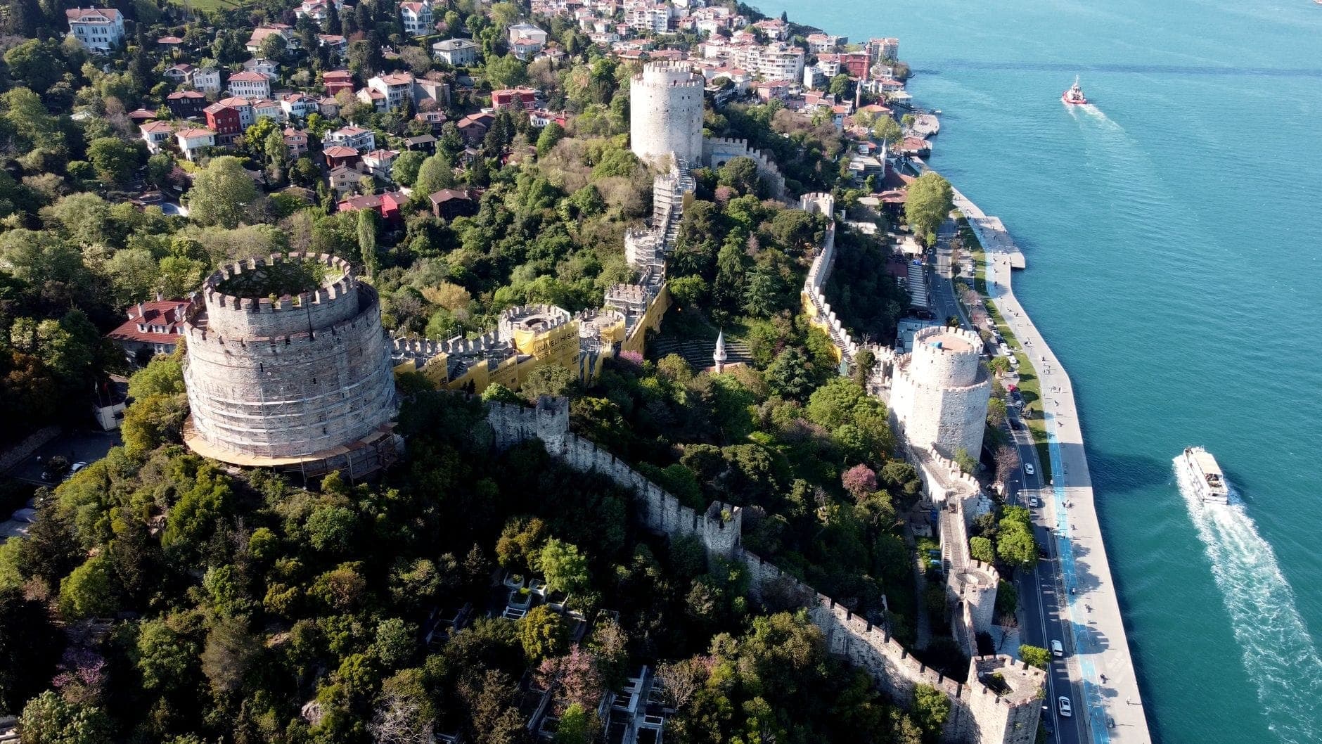 Dramatische Luftaufnahme der Rumeli Festung mit ihren massiven Türmen, historischen Mauern, üppigem Grün und dem angrenzenden Bosporus, im Hintergrund Boote und Stadtgebäude.