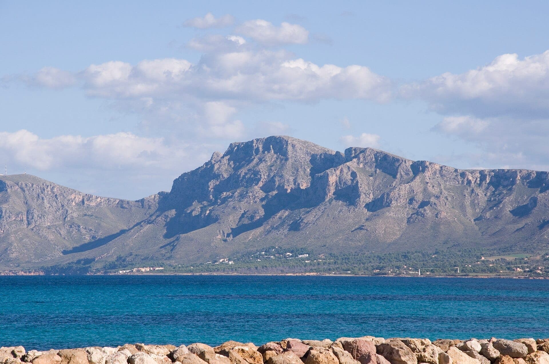 Sa Talaia mountain rising above the green countryside, viewed across blue water under a partly cloudy sky, with rocky shoreline in foreground.