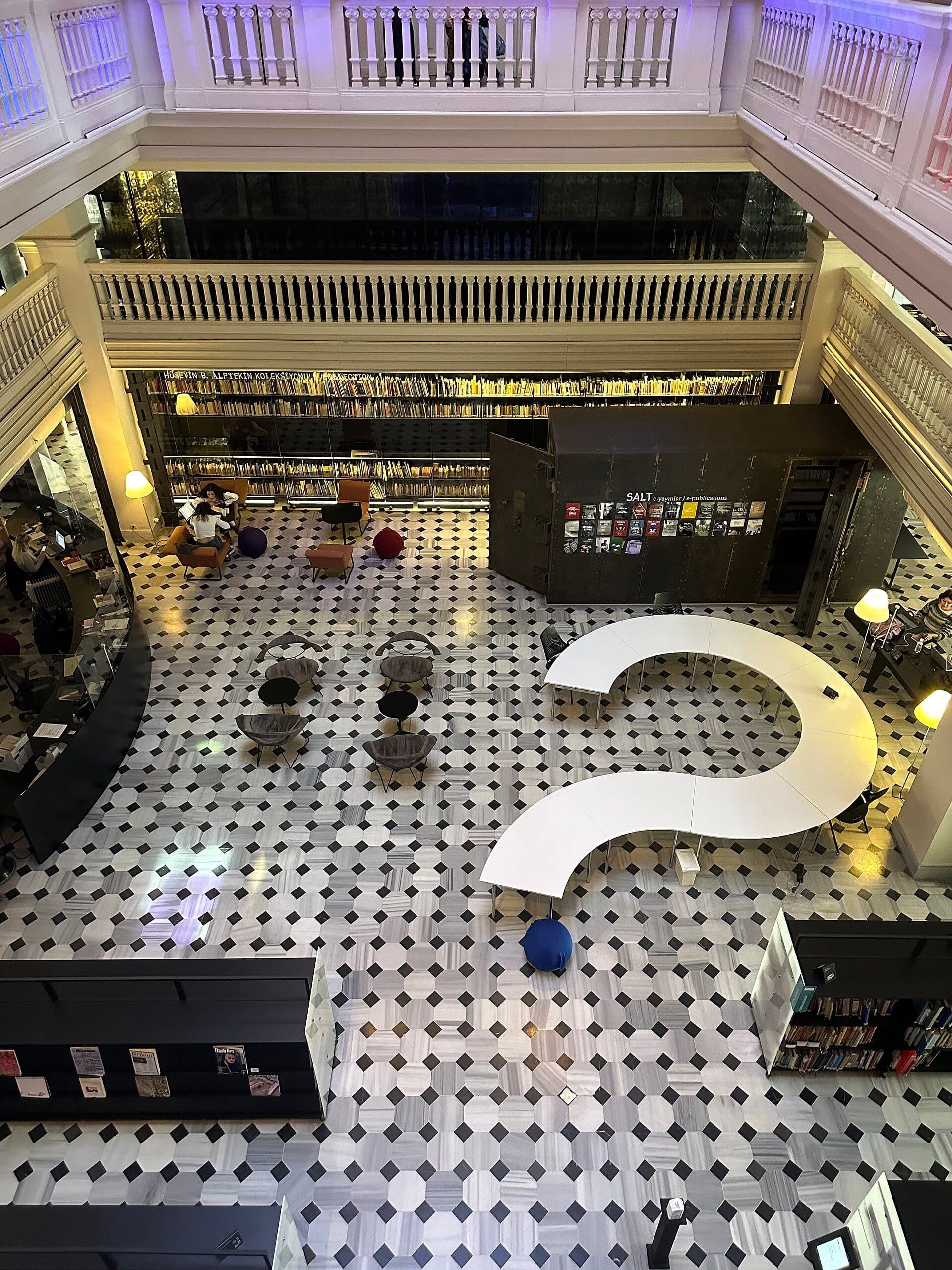 Blick in das luftige Innere von SALT Galata mit Mosaikboden, modernen geschwungenen Tischen, Bücherregalen und gemütlichen Sitzgelegenheiten unter einem großen Zwischengeschoss.