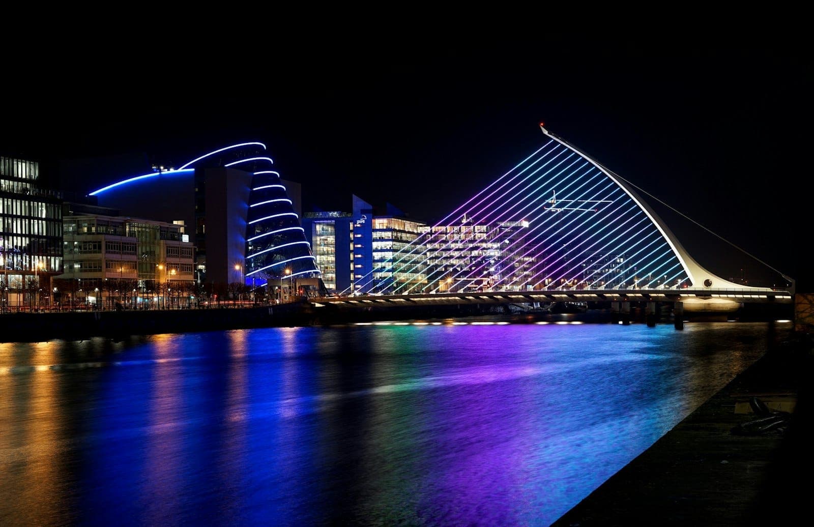 El puente Samuel Beckett iluminado de noche, reflejando luces de colores en el río Liffey con edificios modernos al fondo.