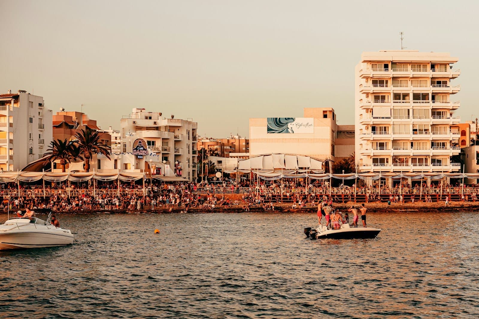 View of San Antonio waterfront at sunset with lively crowds, white resort buildings, palm trees and boats floating on the calm bay.