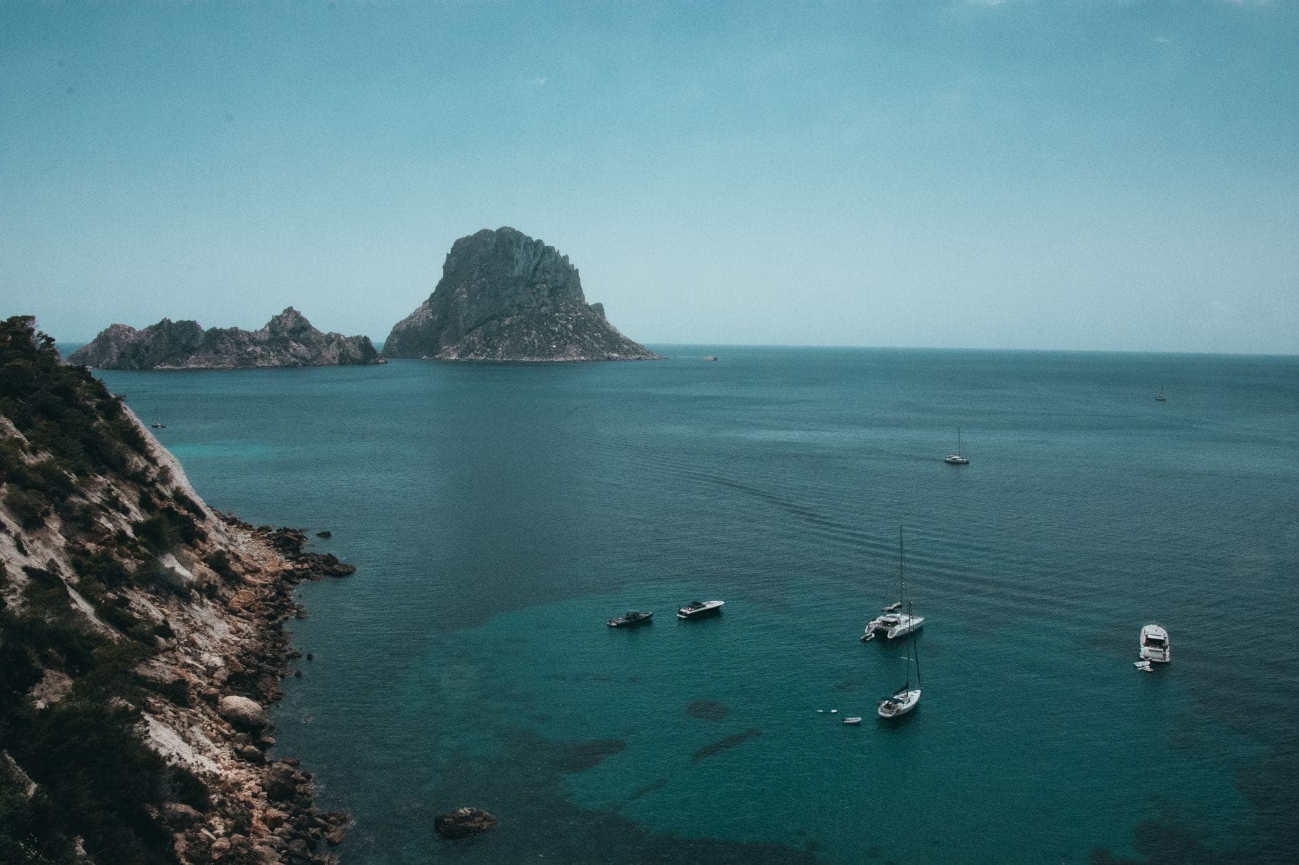 Dramatic view of Es Vedrà islet rising from turquoise sea, with sailboats floating nearby and rugged coastline in the foreground under a blue sky.