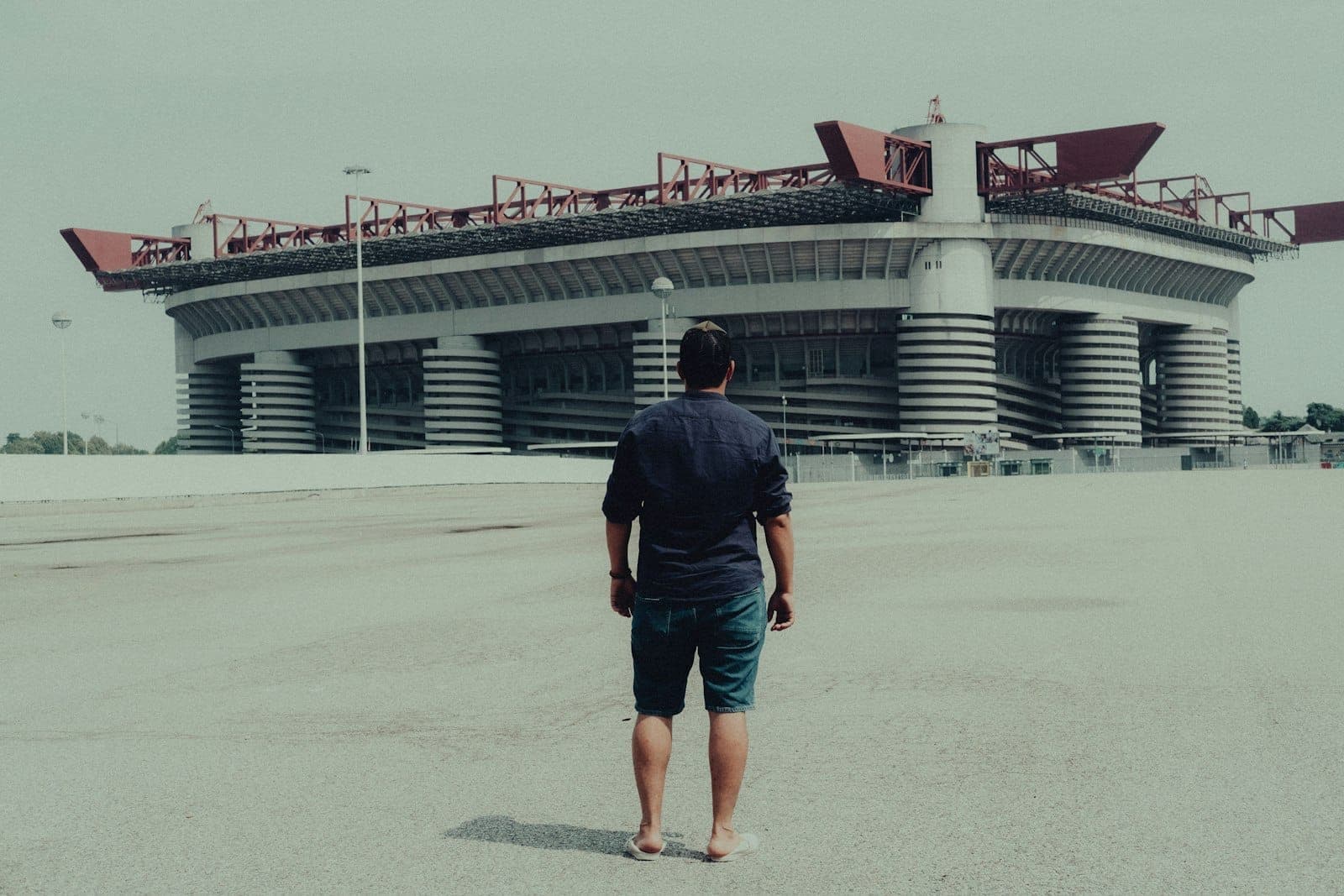 A person stands on an empty plaza in front of the massive San Siro Stadium, with its distinctive red roof and cylindrical columns.