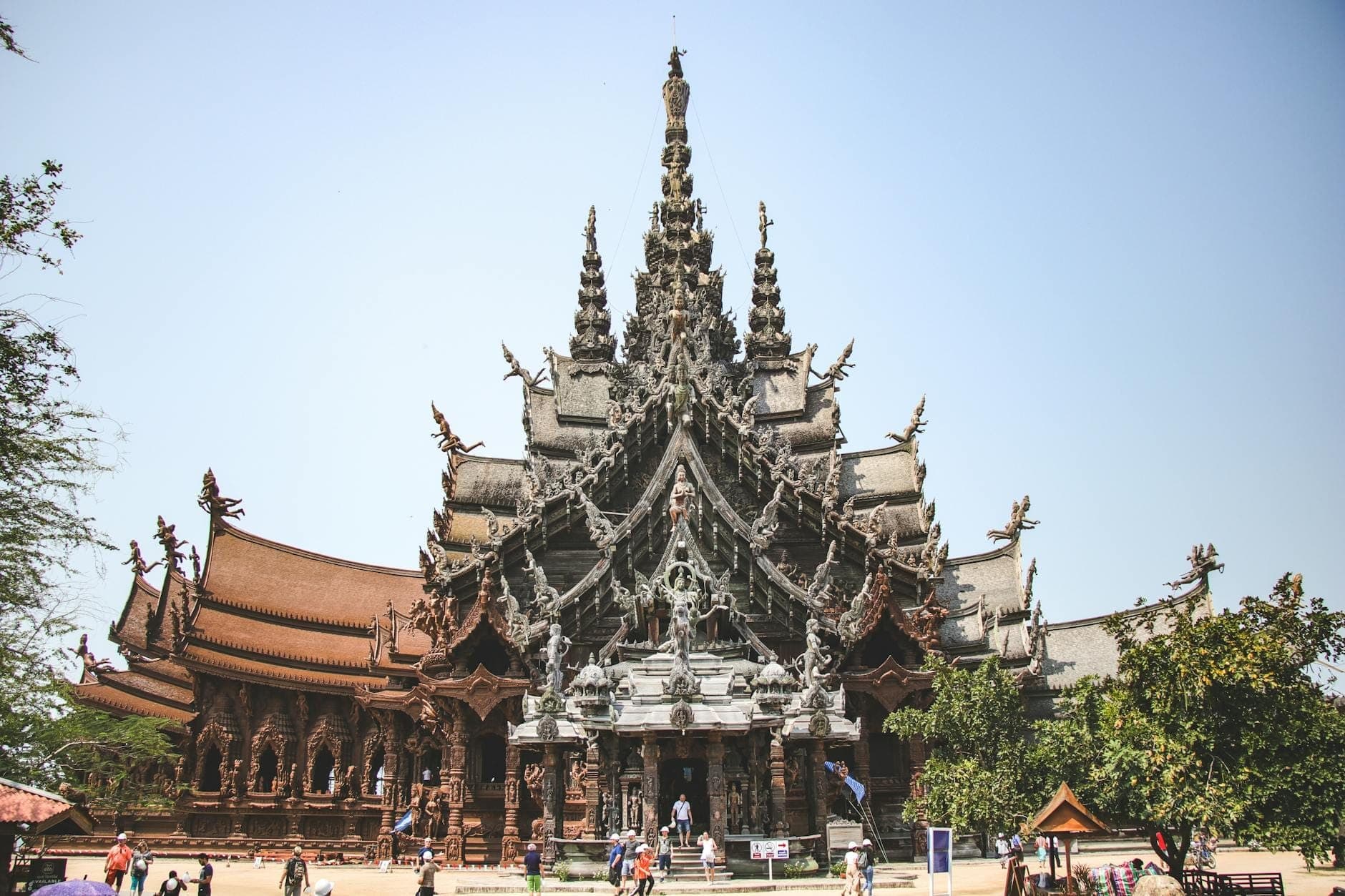 Wide view of the Sanctuary of Truth temple in Pattaya, showcasing its towering wooden spires and detailed carvings under a clear sky with visitors at the entrance.