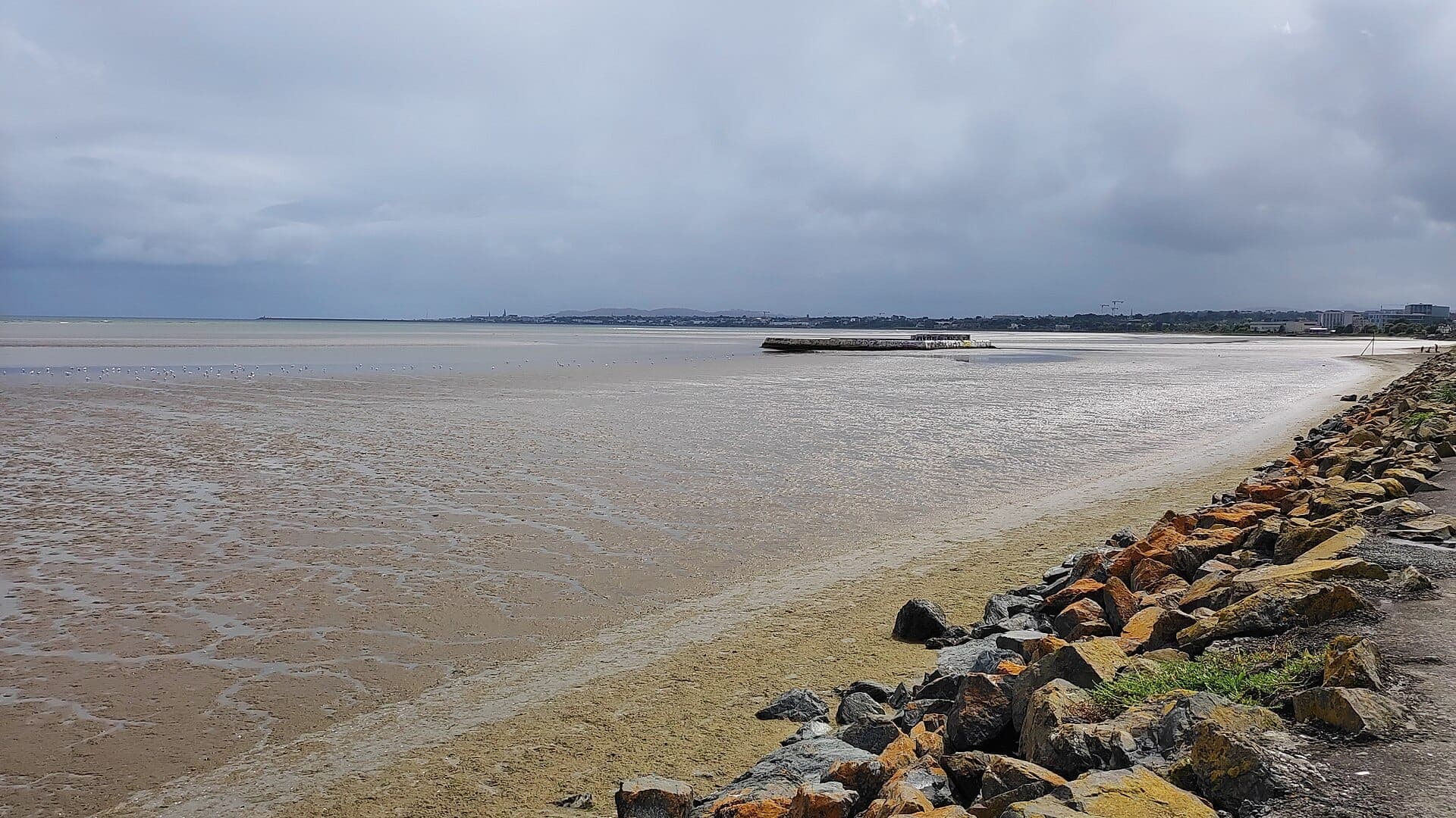 Sandymount Strand bei Ebbe mit weiten Sandflächen, felsiger Küstenlinie, entferntem Pier und bewölkter Skyline der Dublin Bay.