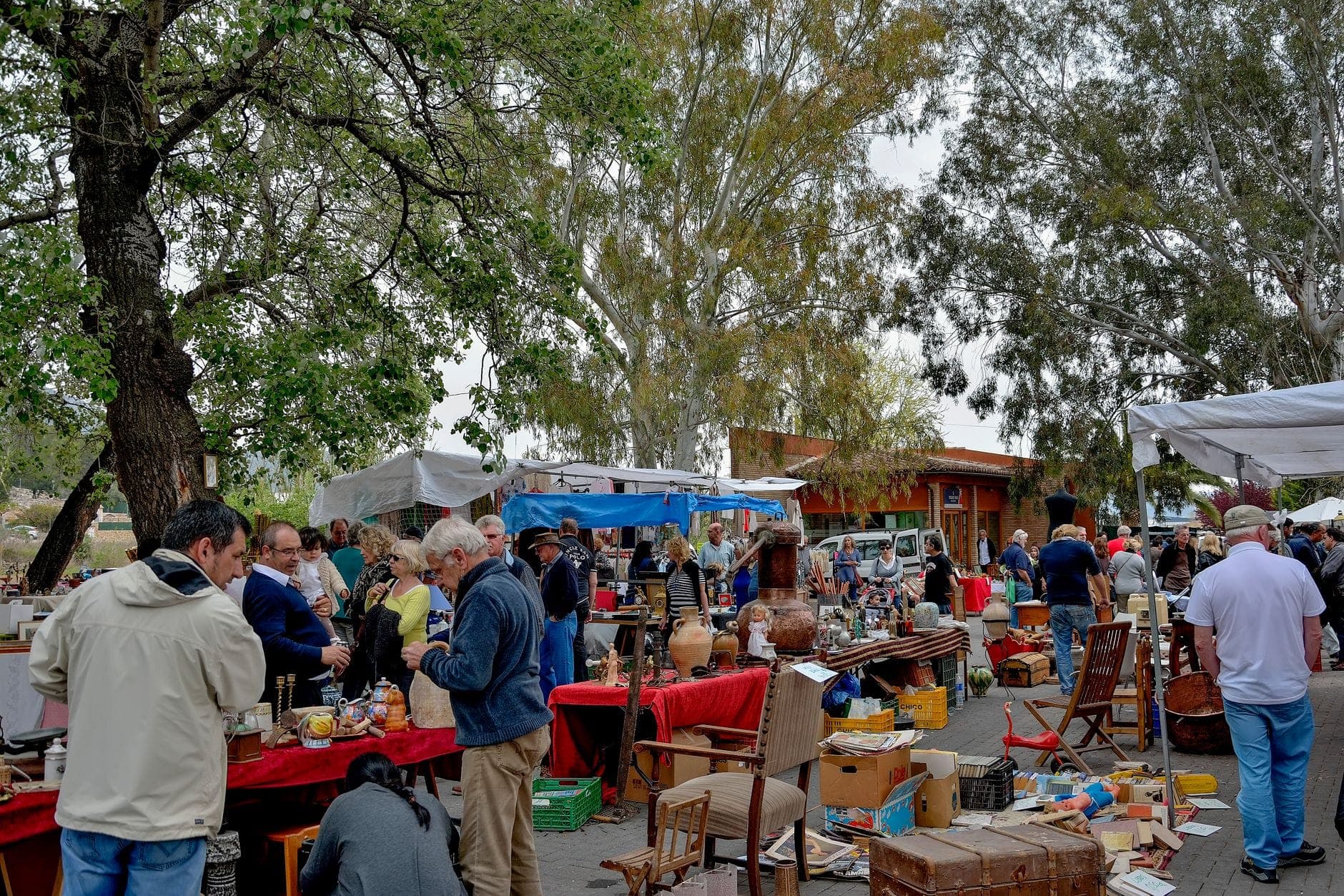 Busy outdoor flea market with people browsing various tables of antiques and vintage goods beneath trees, capturing the bustling local atmosphere of Sant Jordi Flea Market.