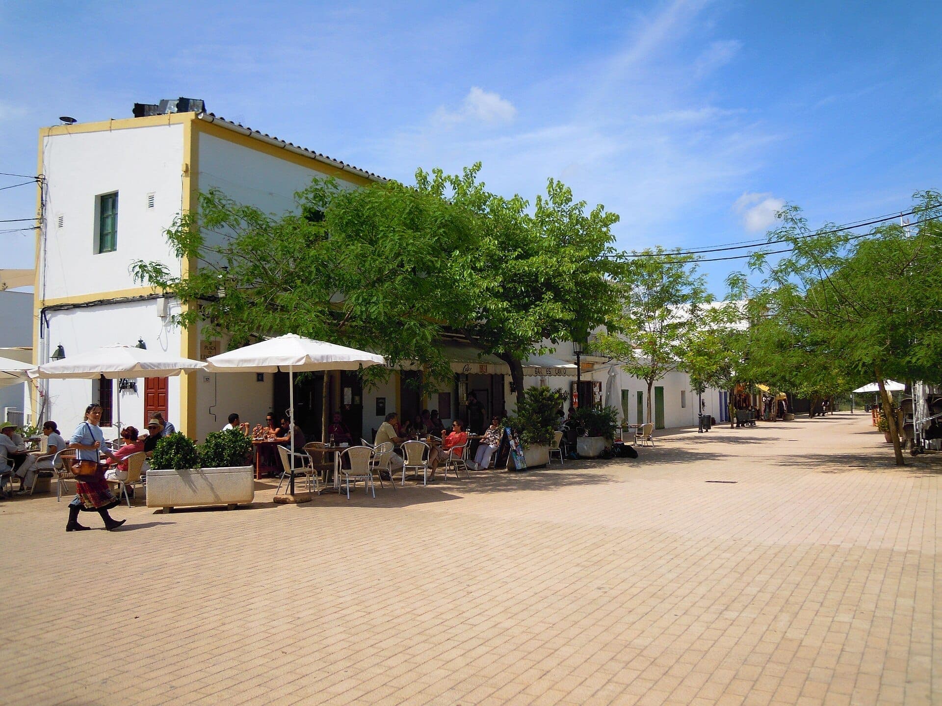 Sunlit village square in Santa Gertrudis de Fruitera, Ibiza, with whitewashed buildings, café umbrellas, trees, and people enjoying outdoor seating.