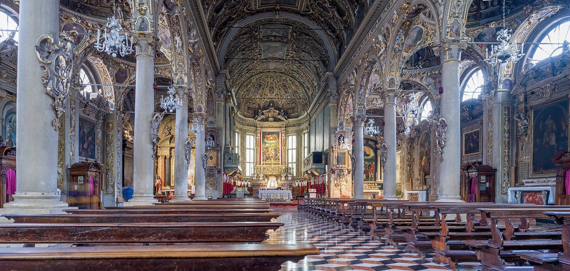 Interior view of Santa Maria delle Grazie in Milan, showing vaulted ceilings, decorative columns, chandeliers, and wooden pews facing the ornate altar.