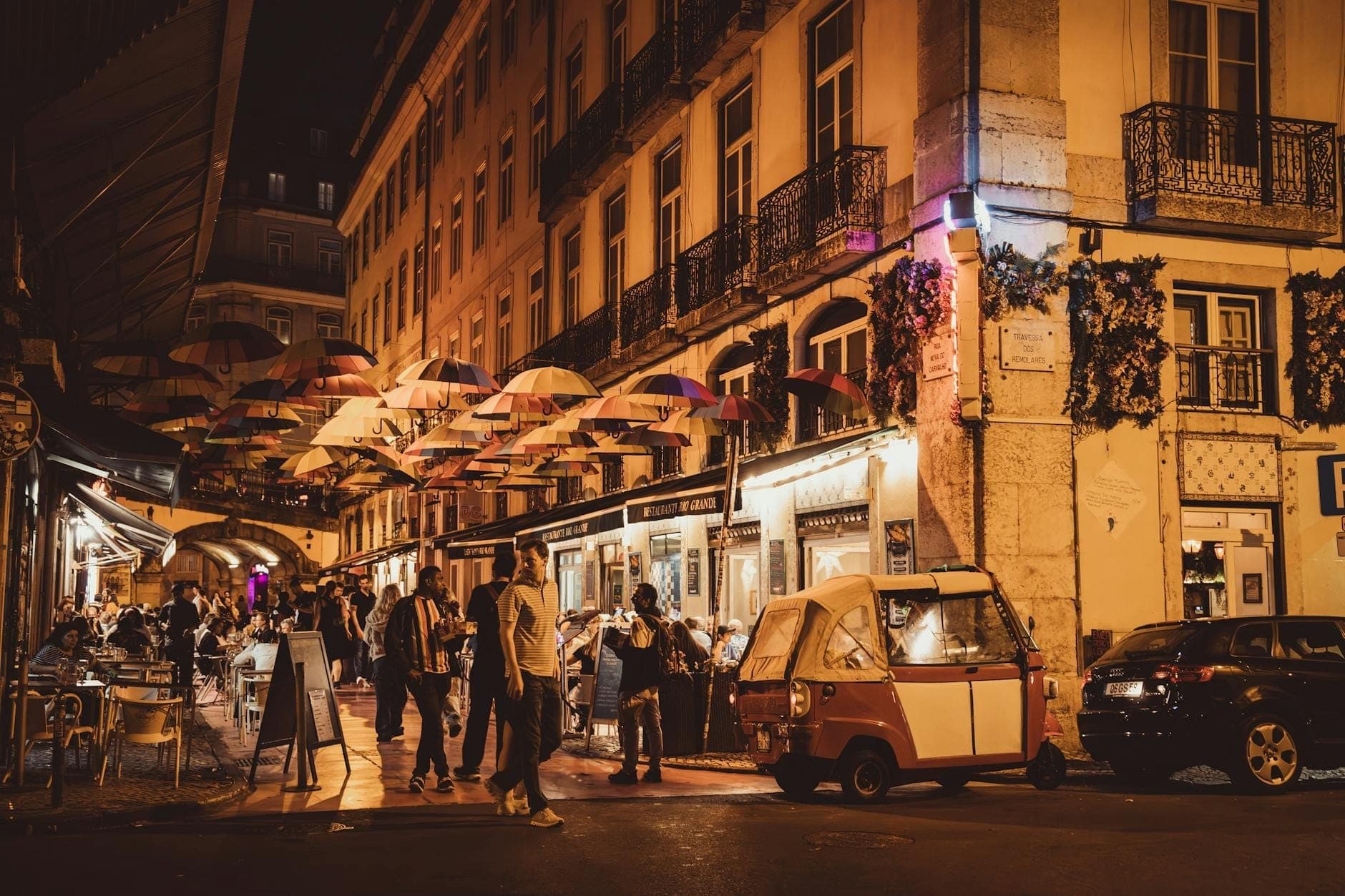 Khung cảnh sôi động về đêm trên đường phố Lisbon với người dân tụ họp, nhà hàng và những chiếc ô đầy màu sắc treo trên đầu, mang đúng không khí lễ hội Santo António.
